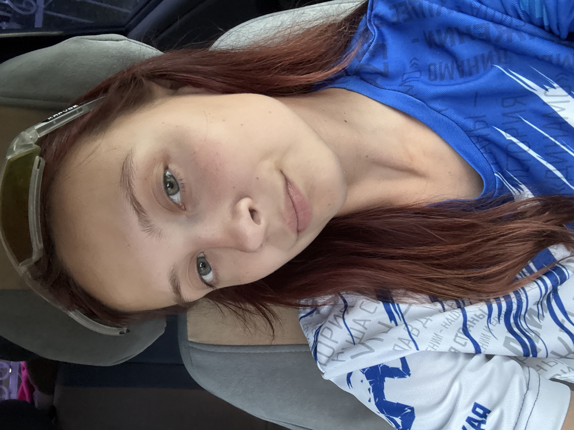 A young woman with reddish-brown hair and green eyes is pictured in a close-up selfie. She is wearing a white and blue sports jersey. The background shows the interior of a vehicle, suggesting she might be a passenger or driver. The scene is indoors, within a car. The woman is looking directly at the camera, and her expression is neutral. Her hair is partially covered by a pair of clear-lensed glasses resting on her forehead. There is no discernible activity or interaction taking place. The setting does not provide any specific clues to pinpoint a location within the city of Chaikovsky, Russia.