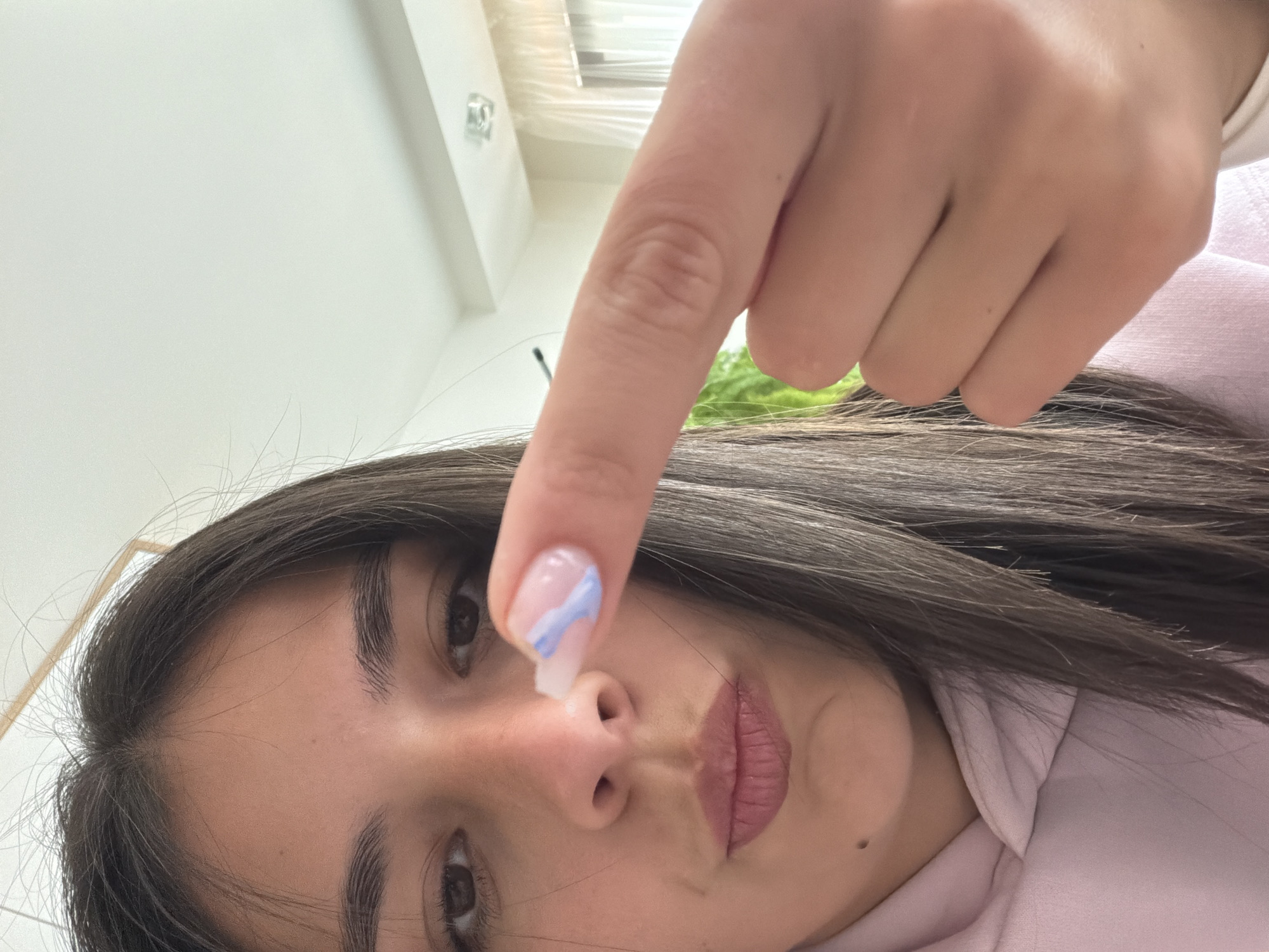 A young woman with brown hair and eyes, wearing a pink hoodie, is holding her index finger towards the camera, near her nose. Her fingernail is painted with a clear base and a light blue stripe design. The background includes a white ceiling and a window with green plants visible outside. There is no indication of the location being Gabrovo, Bulgaria.