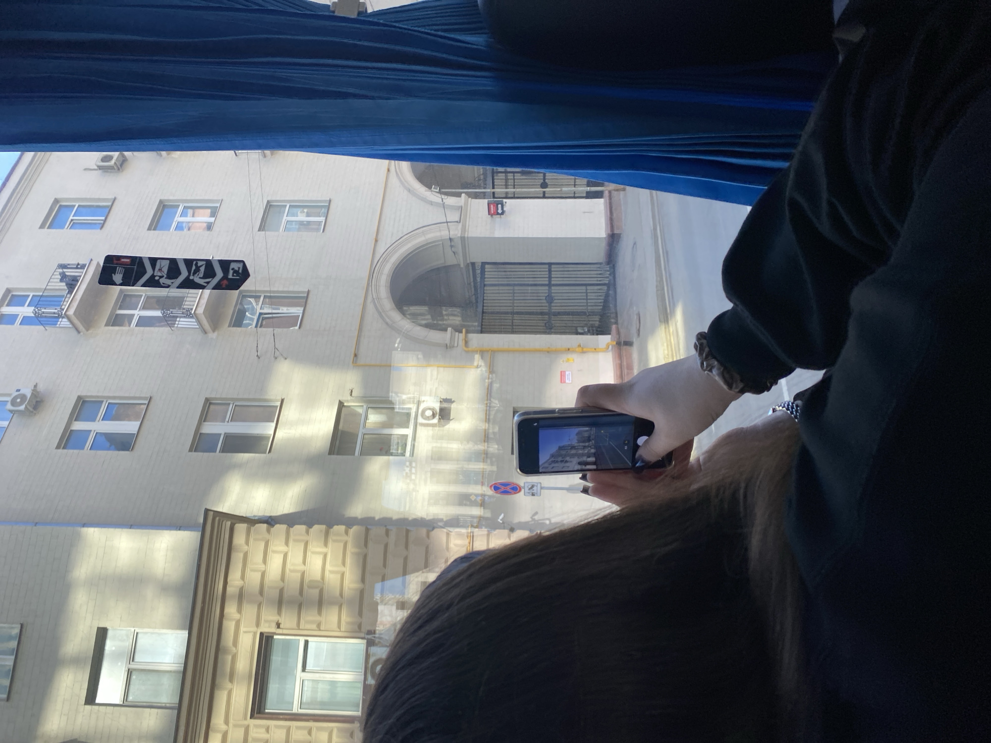 A person in a black sweater is taking a photo of a building through a window, possibly on a bus. The building has several windows, air conditioning units, and a sign with human figures depicting workplace safety instructions. The building also features a large arched gateway. This photograph was taken in the city of Moscow, Russia.