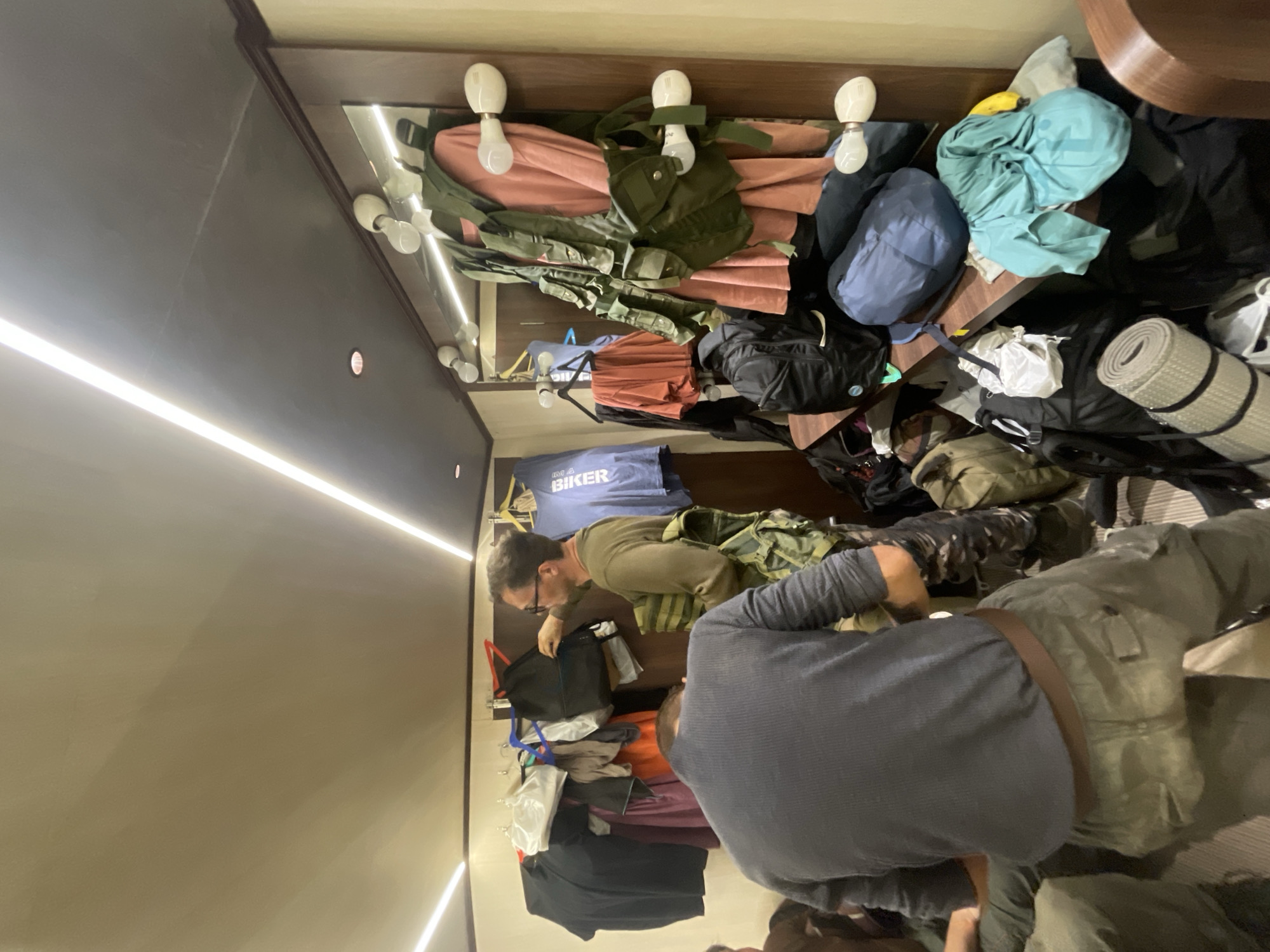 Two men are packing or unpacking items in a small, cluttered room. The room appears to be a storage space or closet. Garments and backpacks hang from hooks on the top of the wall. One man wears a gray shirt. The other man wears glasses and a green long-sleeved shirt.
The location cannot be determined as Mira-Bhayander, India from the image.
