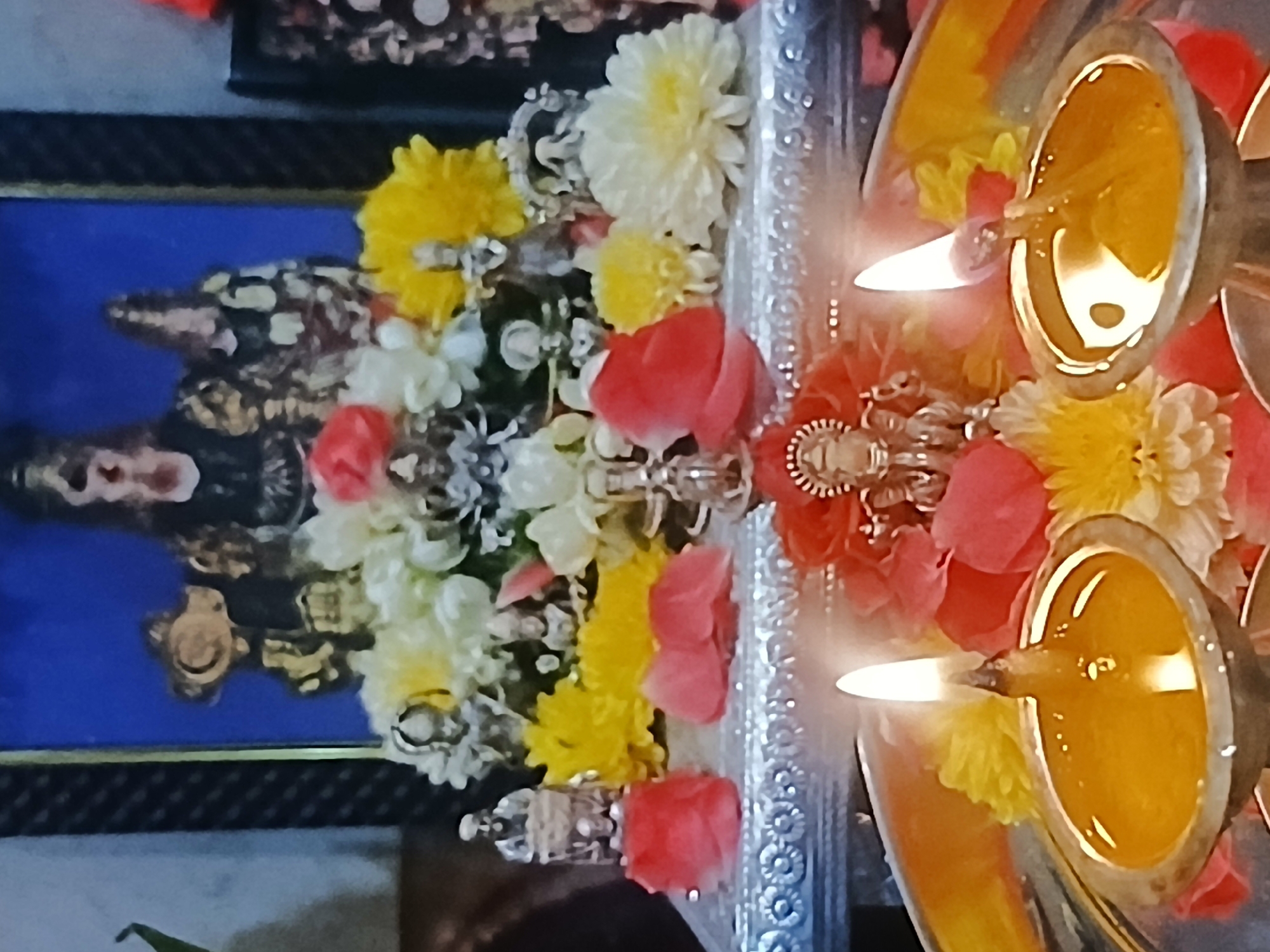 A close-up shot presents a religious display. Two oil lamps with ignited wicks are positioned prominently. Floral arrangements, including white, yellow, and red flowers, are placed around the lamps. Behind the flowers, a framed picture is visible, depicting a figure on a blue background. The scene is set on a silver-colored tray with an ornate edge. Given the imagery, this is likely a Hindu religious setting. The location is Bengaluru, India, based on cultural indicators within the image.