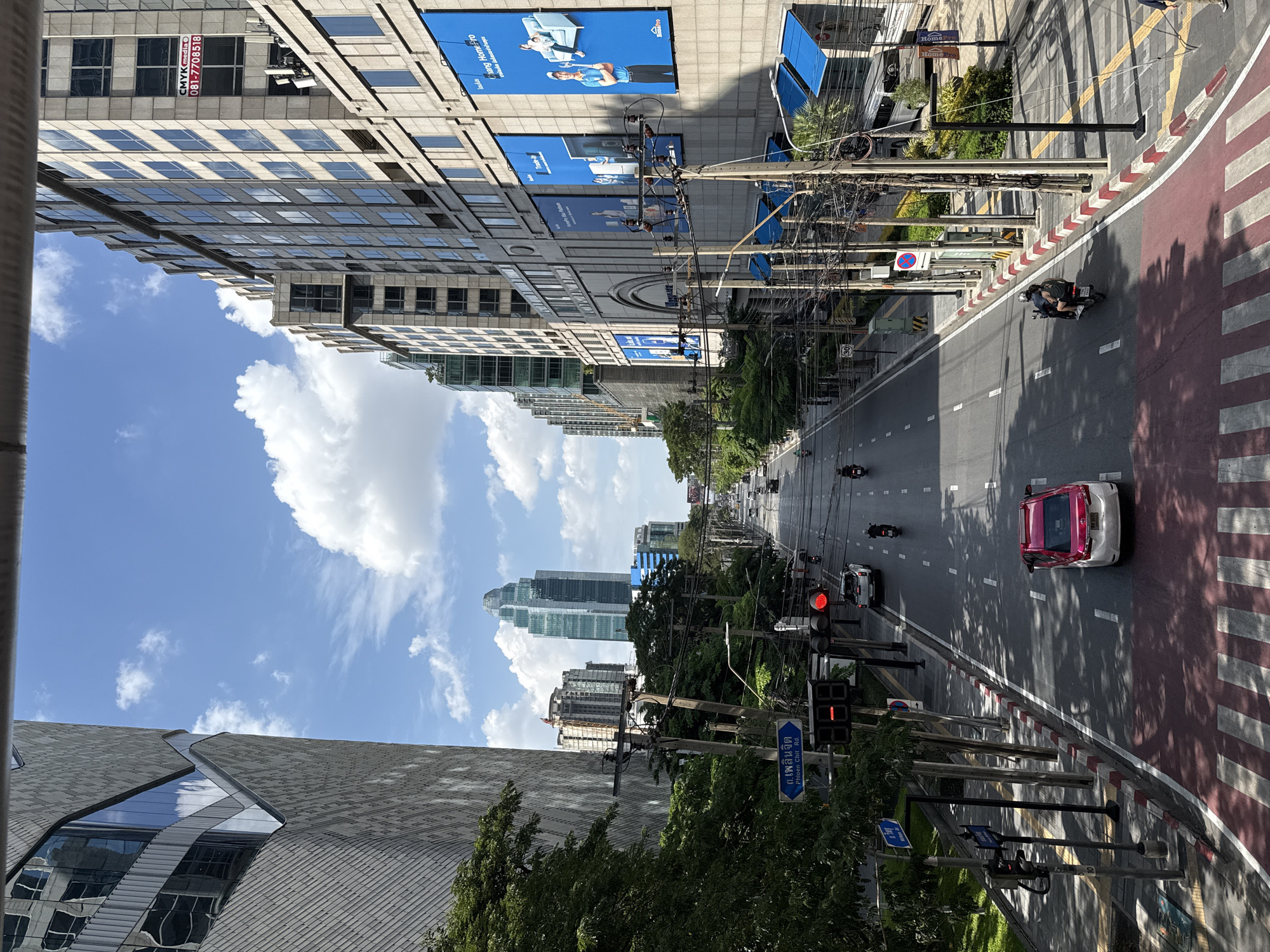 The image depicts a street scene in Bangkok, Thailand, likely in a commercial or business district. A wide, multi-lane road runs through the center, with traffic including cars, motorcycles, and a van. On either side of the road are tall, modern buildings with many windows, some adorned with large blue billboards. Trees line the sidewalks, and numerous electrical wires and poles are strung across the scene, characteristic of urban infrastructure in Bangkok. A pedestrian crosswalk in the foreground marks a transition point. The sky is blue with scattered white clouds, suggesting it is daytime. A sign in Thai script, with English text "Phloen Chit Rd.", is visible on the left side of the street, confirming the location.