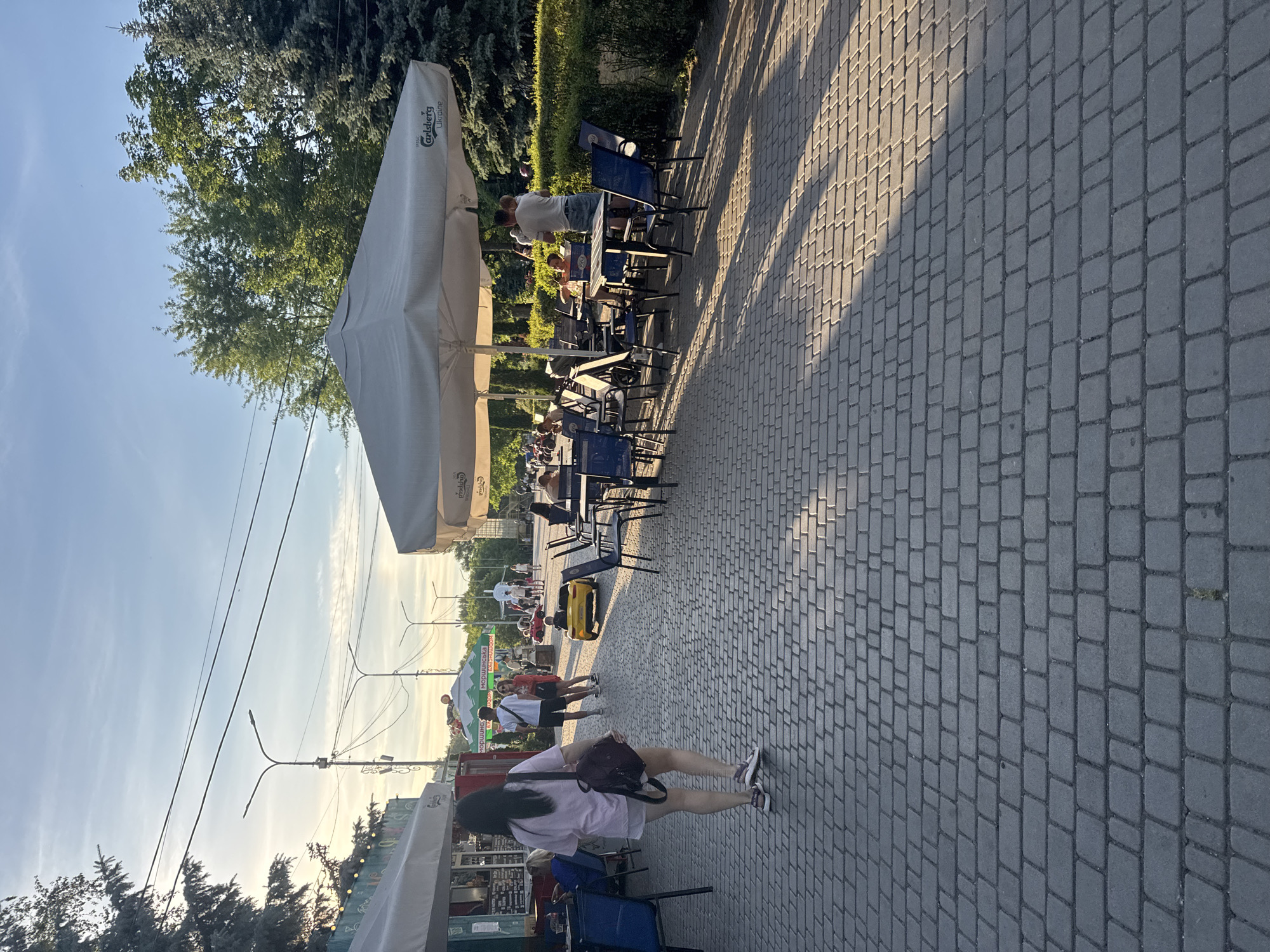 This media file depicts an outdoor scene, likely a street or promenade, featuring a paved walkway lined with tables and chairs. The scene is set under a clear sky, suggesting fair weather, and the lighting indicates it is late afternoon or early evening, given the long shadows cast by the trees and umbrellas.
Several people are present, some sitting at the tables and others walking along the path. A woman in a pink shirt and dark shorts, carrying a backpack, is walking towards the viewer. In the background, more people are visible, including a man standing near a table and a group of people near a green tent. There's also a yellow ride-on toy in the middle ground, suggesting a family-friendly area.
Prominent objects include large, off-white umbrellas providing shade over the seating areas. One umbrella has text on it that reads "Carlsberg Ukraine". There are also streetlights and overhead wires, indicating an urban or semi-urban environment. The walkway is made of interlocking gray pavers.
The overall atmosphere appears relaxed and casual, as people are enjoying the outdoor space, possibly at a cafe or restaurant. No specific event is clearly identifiable, but it seems to be a public area where people gather.