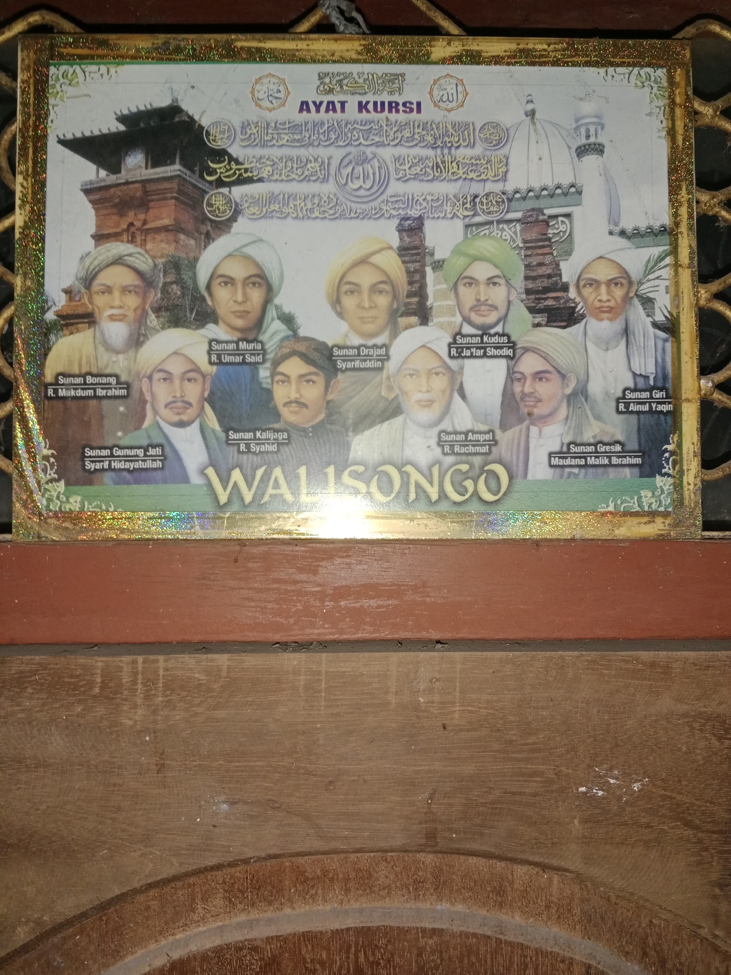 The image shows a framed poster depicting nine figures dressed in traditional Islamic attire, likely historical Islamic scholars. Each figure has their name written below them, with the title "Sunan" followed by their name. The text below the figures reads "WALISONGO" in large, golden, decorative letters. The poster also features Arabic calligraphy and images of what appear to be mosques or religious buildings in the background. The poster is framed with a gold and glittery border. The poster is placed against a wooden surface. The location of Ringinrejo, Indonesia, is not visible or suggested by any elements in the image itself.
