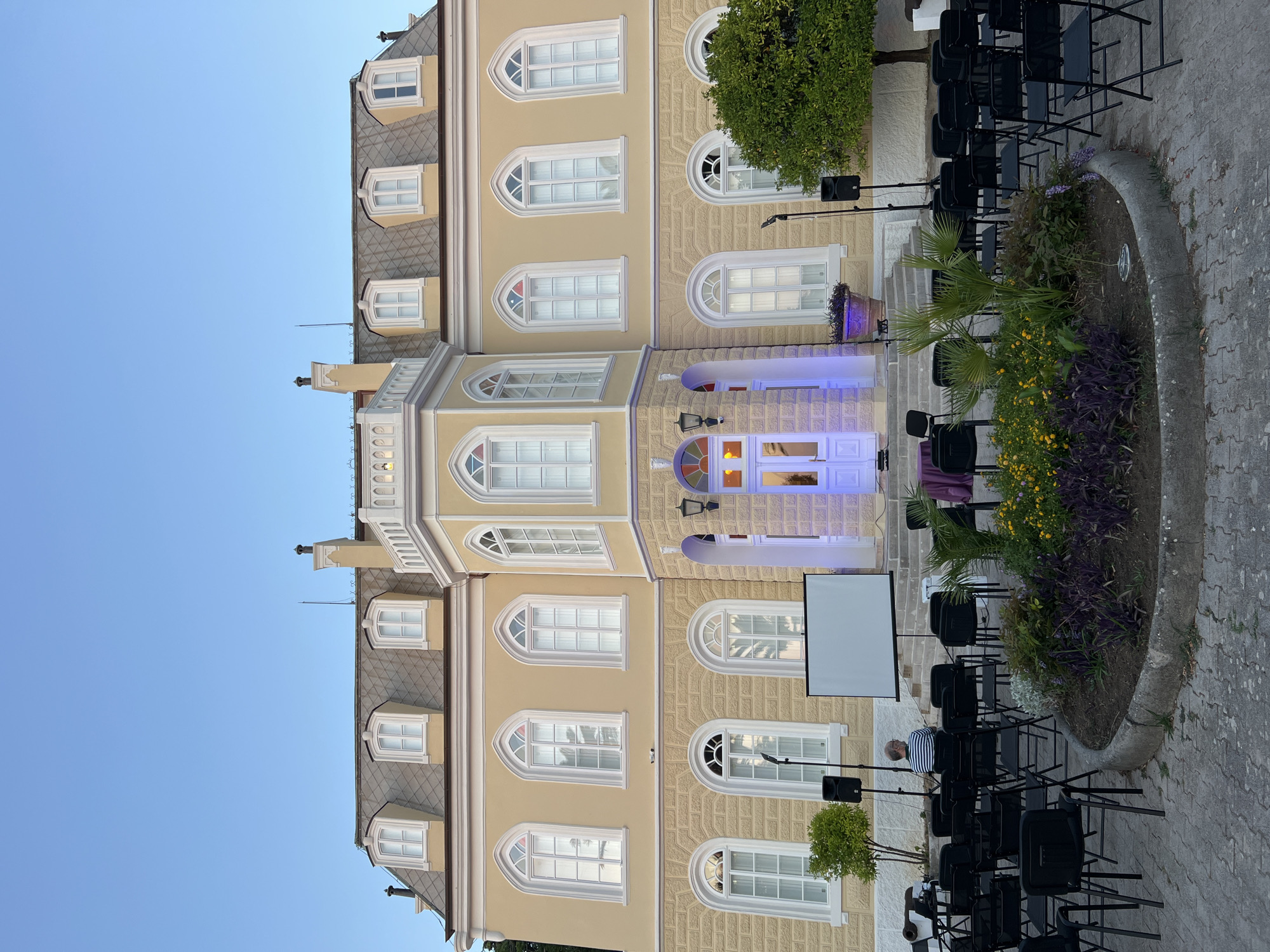 This media file shows an outdoor seating area set up in front of a pale yellow, multi-story building. Rows of black chairs are neatly arranged on a paved surface, facing a white projection screen that is propped up on a stand. A low, curved concrete border surrounds a garden bed filled with lush green plants, purple foliage, and small yellow flowers.
The building features numerous arched windows with white frames, and several have decorative stone trim. The architecture suggests a European style, possibly a hotel or a historic building. One of the windows on the ground floor is lit with blue light, creating a vibrant contrast with the building's facade. To the right of the entrance, a small, leafy green tree stands tall.
There are two black speakers on stands positioned strategically near the seating area, suggesting an event with sound. In the background, a clear blue sky indicates fair weather, and the lighting suggests it is either early morning or late afternoon. The overall scene evokes a sense of anticipation for an outdoor gathering or presentation. The location cues suggest Bar, Montenegro, and the atmosphere is calm and organized, with no visible text in the image.
