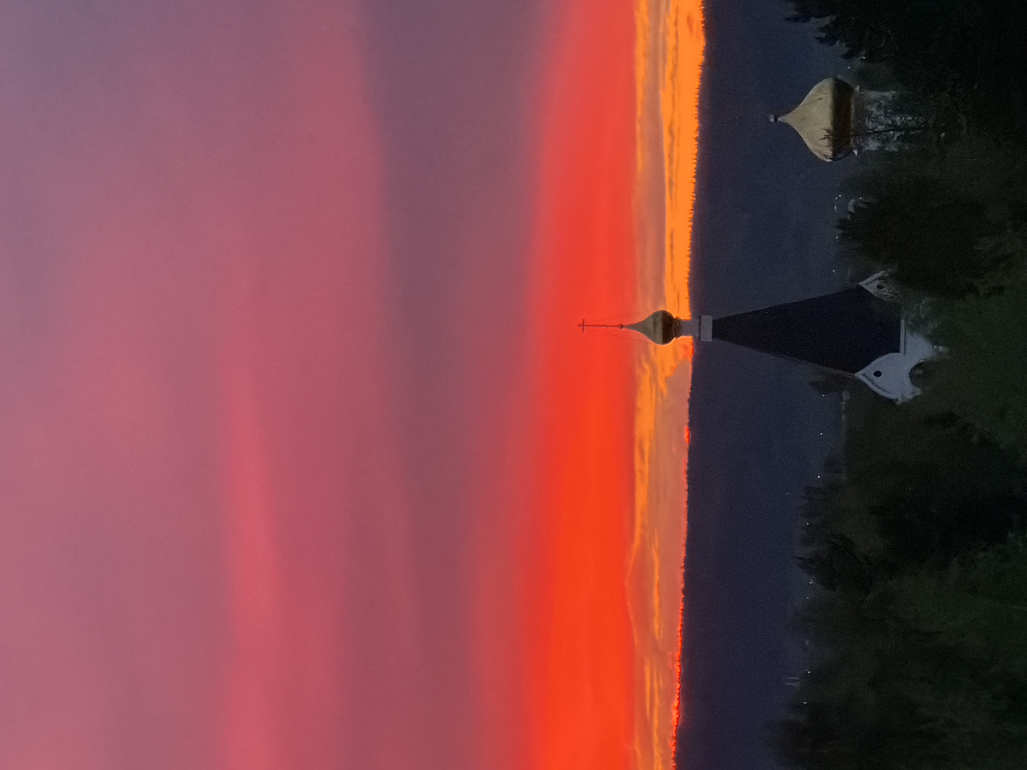 The image captures a vibrant sunset in Ust-Ilimsk, Russia. The sky displays a gradient of colors, transitioning from pink on the left to a brilliant orange in the center. A church with a cross-topped dome stands silhouetted against the sunset. A second, smaller dome is also visible. The foreground is obscured by dark trees, and some lights are visible.