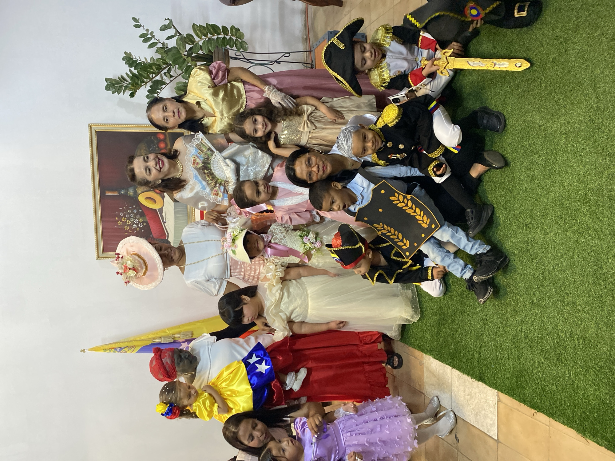 This image captures an indoor group portrait in Tinaquillo, Venezuela, featuring six adults and ten children. The background consists of a plain white wall, a Venezuelan flag on a pole, a framed still-life painting depicting fruit, and a potted green plant on a stand. The floor is tiled light brown on the left and transitions to artificial green turf in the right and foreground sections.
The individuals are dressed in various period-style costumes. In the center-left, an adult female with a dark red headwrap and dark facial paint holds a small child wearing a yellow dress designed with the Venezuelan flag's colors and stars. To her right, an adult female in a white blouse and pink skirt wears a large pink hat adorned with floral elements and holds a small pink bag. Adjacent, another adult female in a shimmering off-white, off-shoulder dress and a pearl necklace holds an open decorative fan. On the far right, an adult female in a golden satin blouse with puffed sleeves and a pink skirt wears white gloves.
In the mid-ground, five young girls stand. From left to right: a girl in a purple frilly dress, a girl in a long cream-colored ruffled dress, a girl in a white lace dress with a pink ribbon and a floral hat, a girl in a shimmering gold dress, and a girl in a light pink dress.
In the foreground, four boys are seated or kneeling on the green turf. From left to right: a boy in a black and gold military-style costume holding a black shield with a gold laurel wreath; a boy in a black military-style jacket with gold epaulets, white pants, medals, and white-painted hair; a boy in a light blue shirt and jeans, holding a black shield with a gold laurel wreath, positioned partially behind an adult female; and a boy in a white, red, and gold military-style jacket, white pants, and a black bicorne hat, holding a gold sword. An adult female wearing glasses is seated among these children, looking towards the camera.
All subjects are generally posed looking towards the camera; most adults and some children exhibit smiles.
