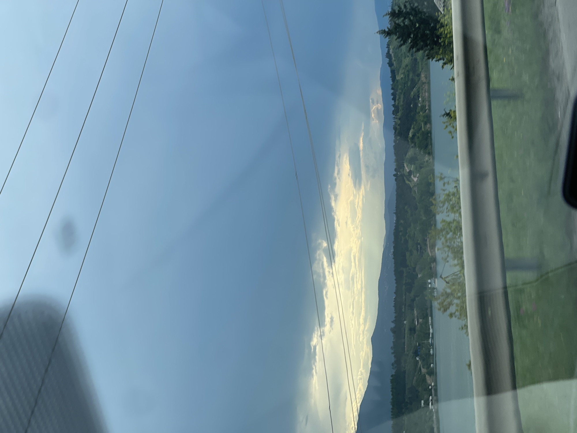 The image shows a landscape with a blue sky, power lines in the upper left, and a view of mountains and a body of water. There are clouds with visible sunbeams. The image appears to be taken from inside a vehicle, as part of the window frame and a guardrail are visible. The location cannot be determined to be Tbilisi, Georgia from the image alone.