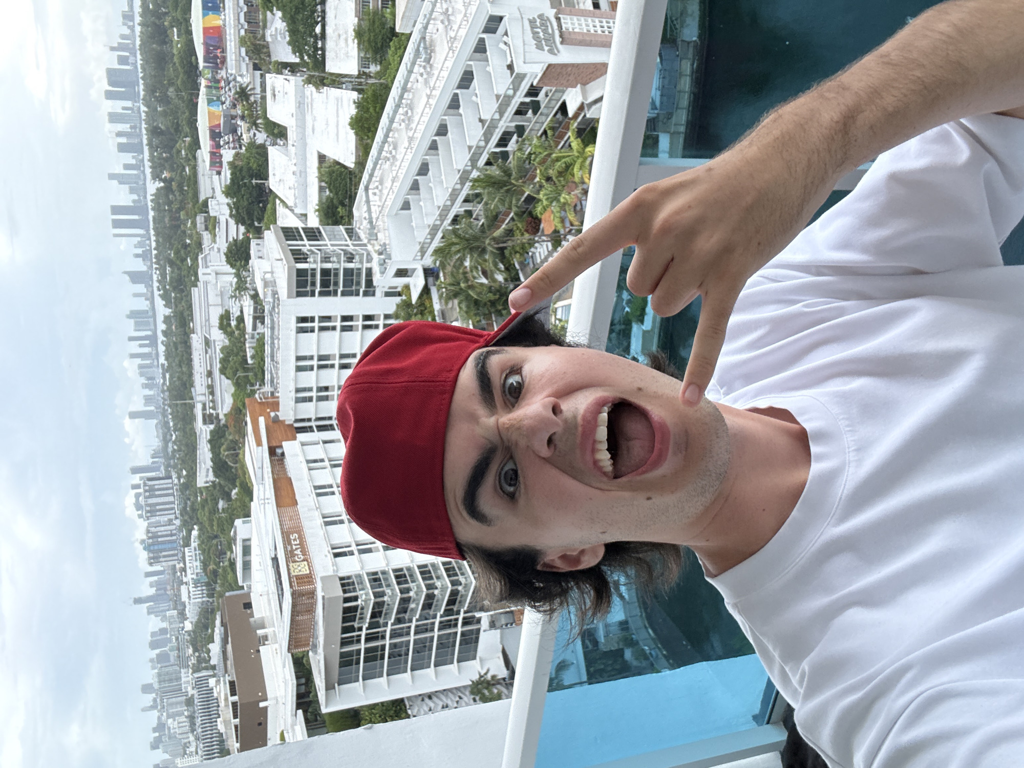 A man wearing a red baseball cap and a white t-shirt is taking a selfie on a balcony. He is making a "rock on" hand gesture and has his mouth open wide, eyes wide. He is standing on a balcony with a blue railing. In the background, there is a view of Miami Beach, Florida. The "Gates" building sign is visible in the background. The weather appears to be overcast.