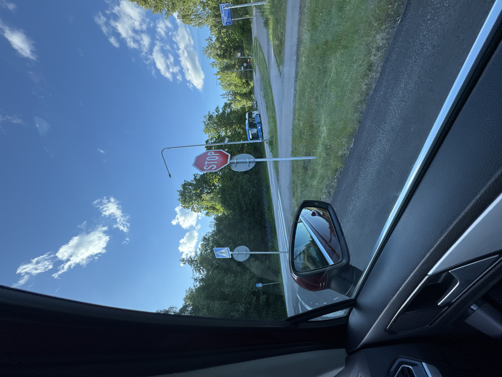 This is a photo taken from inside a car, looking out towards the right side. The image is oriented such that the sky is at the top and the road is in the middle, with trees and greenery lining the side of the road. The photo is taken from the passenger side of the car, as indicated by the side-view mirror visible in the foreground.
In the scene, there are several road signs. The most prominent is a red octagonal stop sign. Further down the road, there are blue square signs with white pictograms, likely indicating pedestrian crossings or other traffic information. A blue bus with white lettering is visible driving away from the camera on the road. The sky is a clear blue with scattered white clouds, suggesting it is daytime. The weather appears to be pleasant and sunny. The presence of lush green trees indicates a natural or suburban setting.
The activity appears to be a car journey, with the camera capturing a view of the passing scenery. There are no people clearly visible in the image, though a reflection in the side-view mirror might show a glimpse of someone. The overall mood is calm and observational, typical of a snapshot taken during travel. The visible text on the stop sign reads "STOP".