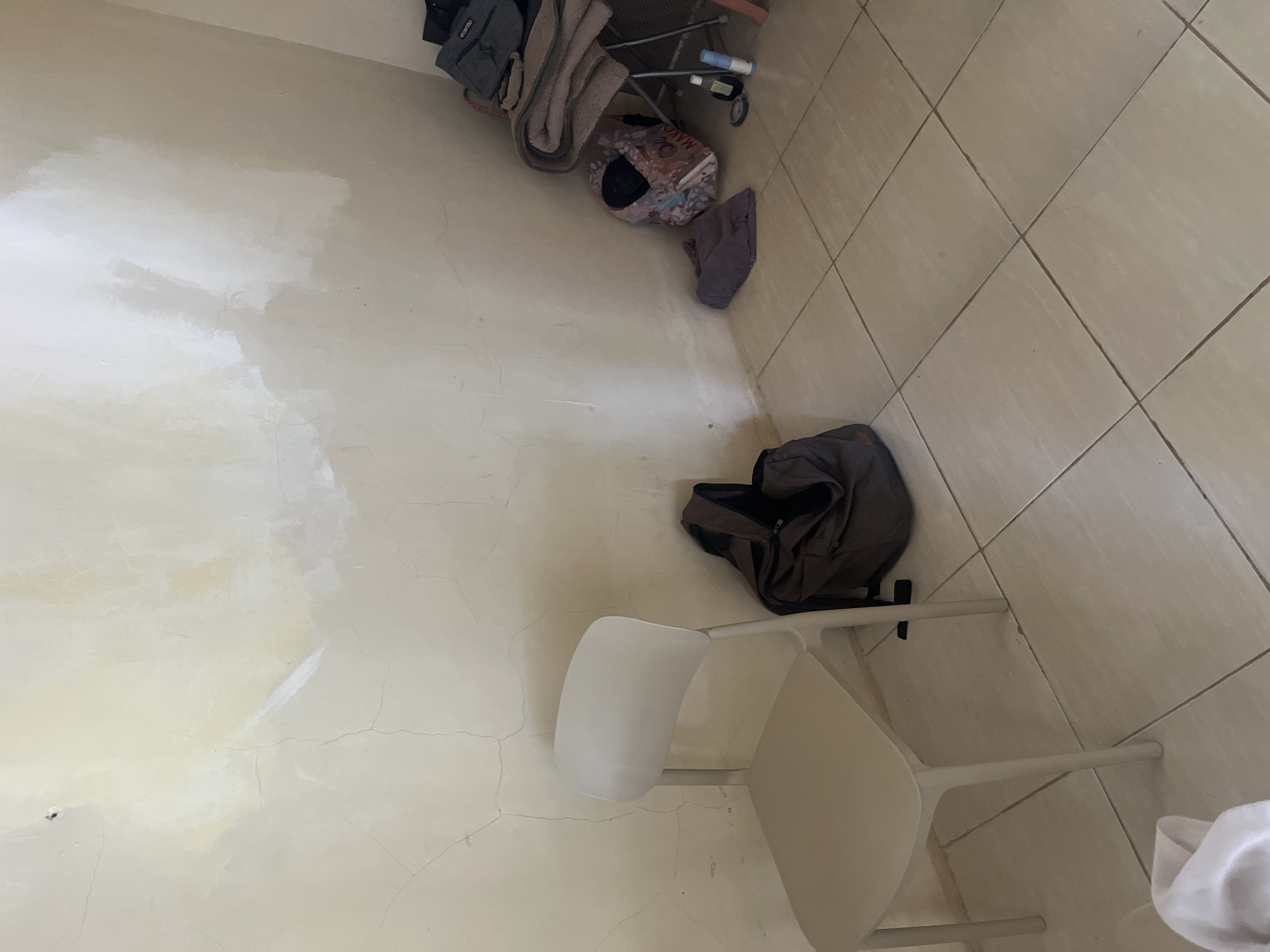 The image depicts an interior scene, likely a room, with light-colored tiled flooring. A plain, light-colored plastic chair stands prominently on the left side of the frame. Adjacent to the chair, on the floor, is a dark-colored duffel bag. Further to the right, against the wall, are several items including a folded set of towels, a patterned tote bag, a folded piece of purple fabric, and a couple of small bottles, possibly toiletries. The wall behind these objects appears to be textured and shows signs of wear, with patches of lighter paint or primer applied in some areas, suggesting a work-in-progress or a state of disrepair. The lighting suggests an indoor environment with some natural light potentially entering from an unseen source. The scene conveys a sense of casual placement of personal belongings. This image was taken in Cirahayu, Indonesia.