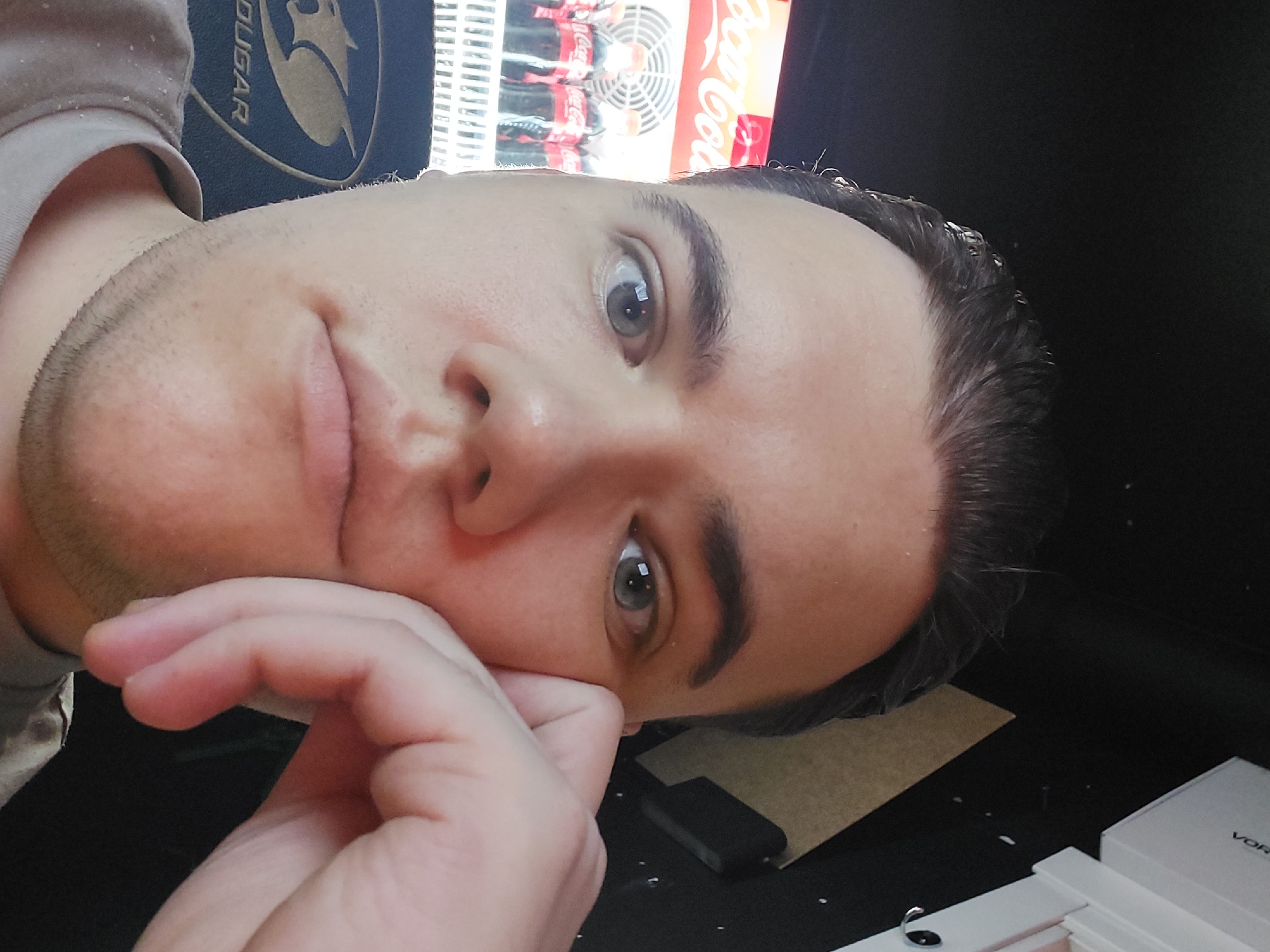A person is resting their face on their hand. The person is wearing a shirt with a "COUGAR" logo on it. A Coca-Cola refrigerator is in the background. Also in the background are packaged soft drinks. The person has light-colored eyes and short dark hair. A smartphone is on a table next to a white box. Given the Cyrillic script of the "Coca-Cola" branding and the location specification of Tashkent, Uzbekistan, it is likely that this scene is taking place in that city or a nearby location.