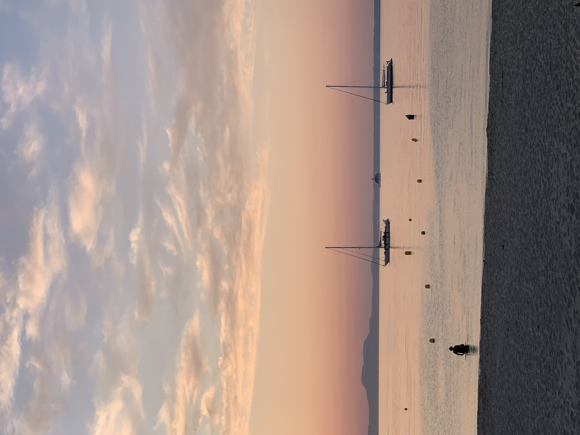 This image depicts a serene sunset scene at the beach in Palmanova, Spain. The sky is a gradient of soft pinks and oranges, with wispy clouds streaked across it. The calm sea reflects the warm hues of the sky. In the foreground, a sandy beach stretches across the bottom of the frame.
In the water, a person is partially submerged, facing away from the camera, appearing to be standing or wading. Several yellow buoys are dotted across the water's surface, marking an area or perhaps part of a swimming zone.
Further out in the sea, two sailboats are anchored. The one on the right is closer and shows a white hull and sails, with a few figures visible on its deck. The sailboat on the left is smaller and appears to have its sails down. In the distance, a larger white vessel, possibly a yacht, is visible on the horizon, along with a faint outline of mountains or hills on the far left of the frame. The overall atmosphere is peaceful and tranquil, characteristic of a sunset by the sea.