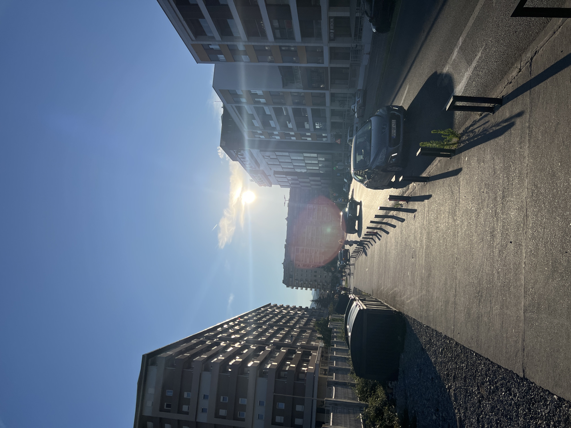 This image captures a street scene in Podgorica, Montenegro, bathed in the warm light of the setting sun. The perspective is from a sidewalk looking down a street lined with modern apartment buildings on both sides. The sun is low in the sky, creating long shadows and a lens flare effect across the scene.
On the left, a tall apartment building with numerous balconies and windows dominates the view. Adjacent to it is a sidewalk with a row of dark, rectangular bins and a gravelly area with some sparse vegetation.
The street itself has two lanes of traffic. Several cars are visible, including a dark gray Peugeot in the foreground, its license plate showing "PV-BF 411". Another silver car is further down the road, and other vehicles are present. Pedestrians are walking on the sidewalk on the right side of the street. The buildings on the right are also modern apartment complexes, with balconies and large windows. One building has a sign that reads "LUCA MEDICAL".
The sky is a clear blue with a hint of a contrail from an airplane. The overall impression is a typical urban environment during the golden hour, suggesting a peaceful evening.