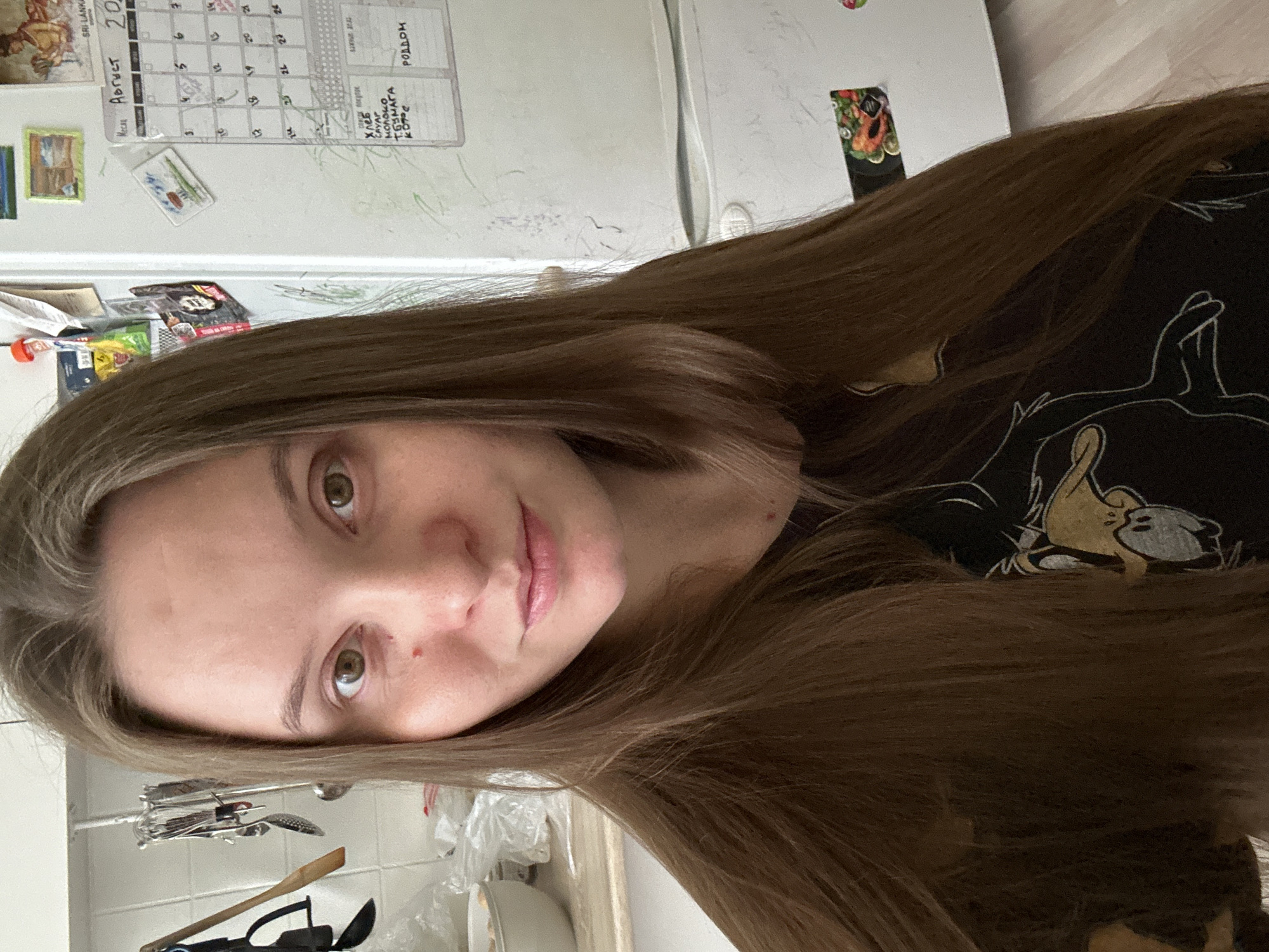 A medium shot shows a young woman with long brown hair and hazel eyes looking directly at the camera. She has a light complexion with some redness on her cheeks and a small red mark on her right cheek. She is wearing a black t-shirt with a cartoon character printed on it.
The background suggests a kitchen environment. To the left, there is a white tiled wall with kitchen utensils hanging on a rack. A white bowl is visible on a counter. To the right, a white refrigerator is present with a calendar and various notes attached to it. The calendar shows the month of August. There are also some drawings and scribbles on the refrigerator door. The overall setting appears to be indoors, possibly a home kitchen.
The location is identified as Noviy, Russia.