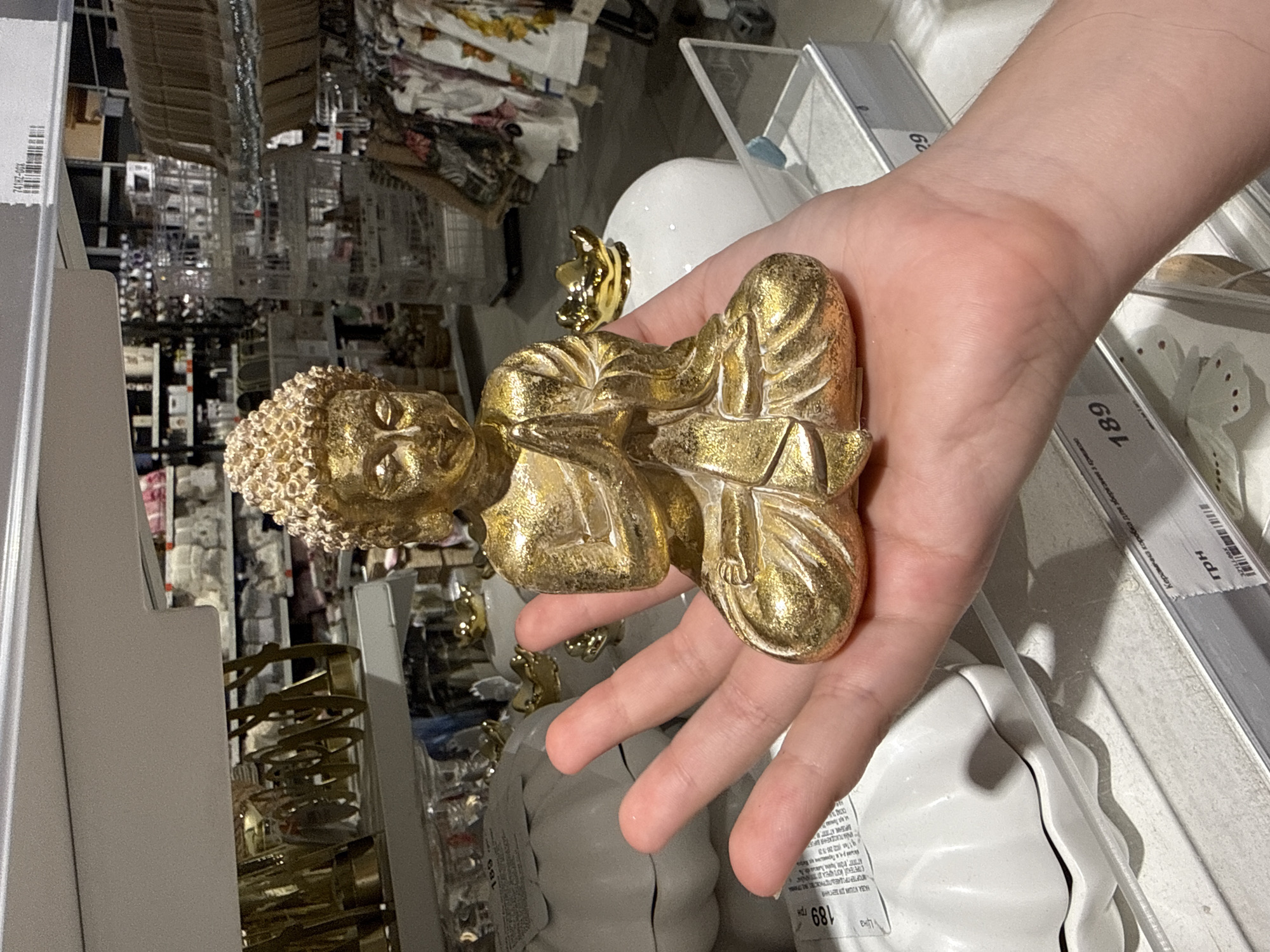 A person's hand holds a small, golden, seated Buddha figurine. The figurine is detailed, with intricate folds in the robe and a textured, curly headdress. A crown-like element rests on top of the headdress. The hand is positioned in a way that suggests the figurine is being presented or examined.
The background is blurry but appears to be a retail environment, possibly a home goods or gift store. Shelves filled with various items are visible, including what look like fabrics or textiles with floral patterns, and other decorative objects. There are also price tags and labels in the background, some with Cyrillic text. One visible price tag reads "189 грн" (189 Hryvnia), indicating the currency of Ukraine. Other visible text includes "АПОРТЕРПРОДАЕАЛІСТРИОХСТВО", "НАЗВА КОШИЯ ДЛЯ ДЕГАННЯ", and "Керамічна коробка для эберезня з грицко".
The lighting suggests an indoor setting, typical of a store. The overall impression is a casual moment of browsing or purchasing in a shop. The exact time of day and weather are not discernible from the image.