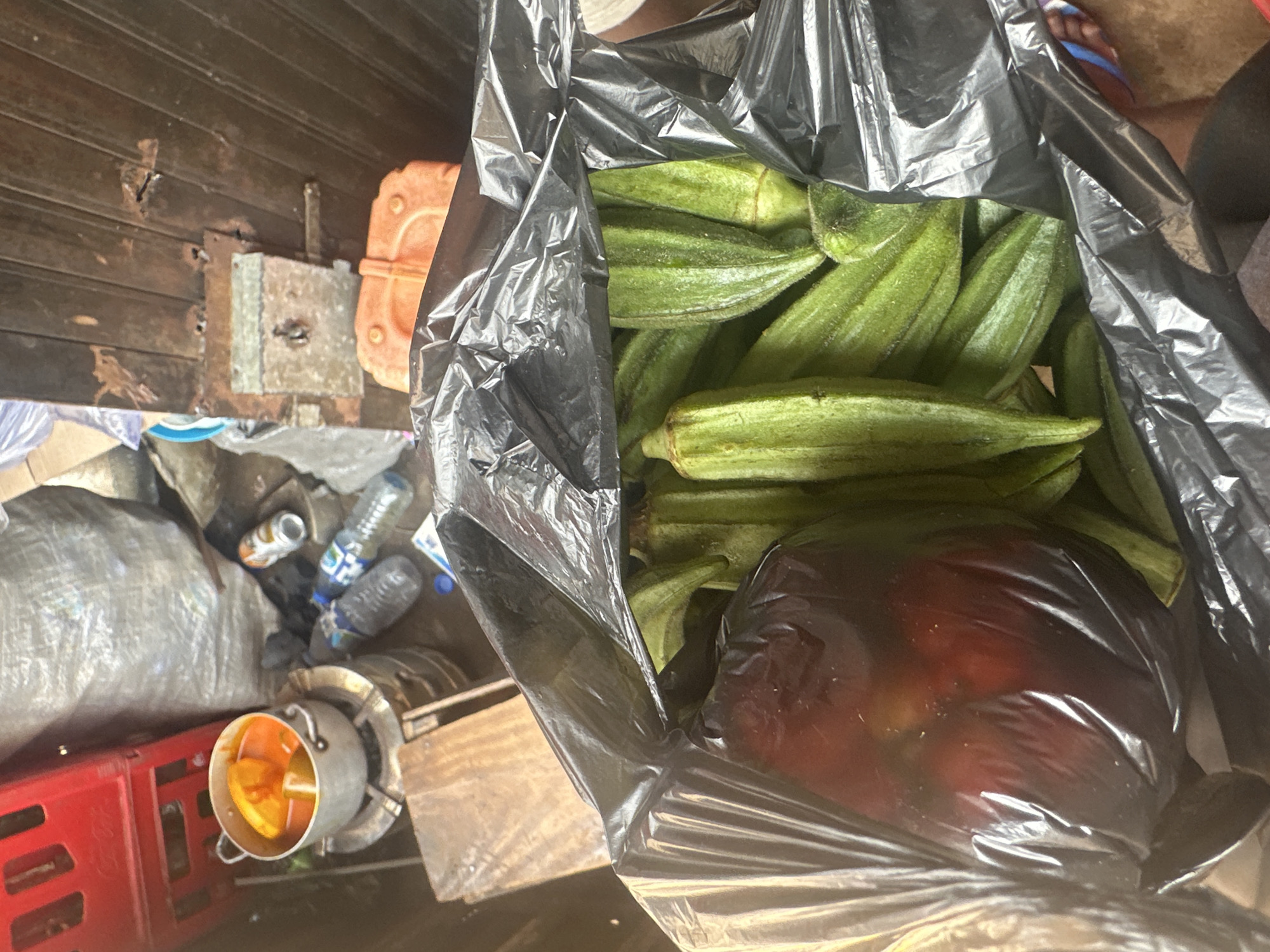 A top-down, medium shot captures a black plastic bag filled with okra in a cluttered outdoor or semi-outdoor environment. The okra pods are bright green and have a ribbed texture. To the left of the okra is a large, dark red object, possibly a fruit or vegetable, also wrapped in black plastic. In the background, various objects suggest a busy marketplace or storage area: a red crate, a silver pot with food on a burner, several plastic bottles, a large grey bag, and an orange container. A wooden surface is visible in the lower left, and part of a wooden door with a metal lock is in the upper right. The overall scene conveys a sense of everyday activity and perhaps the preparation of a meal.