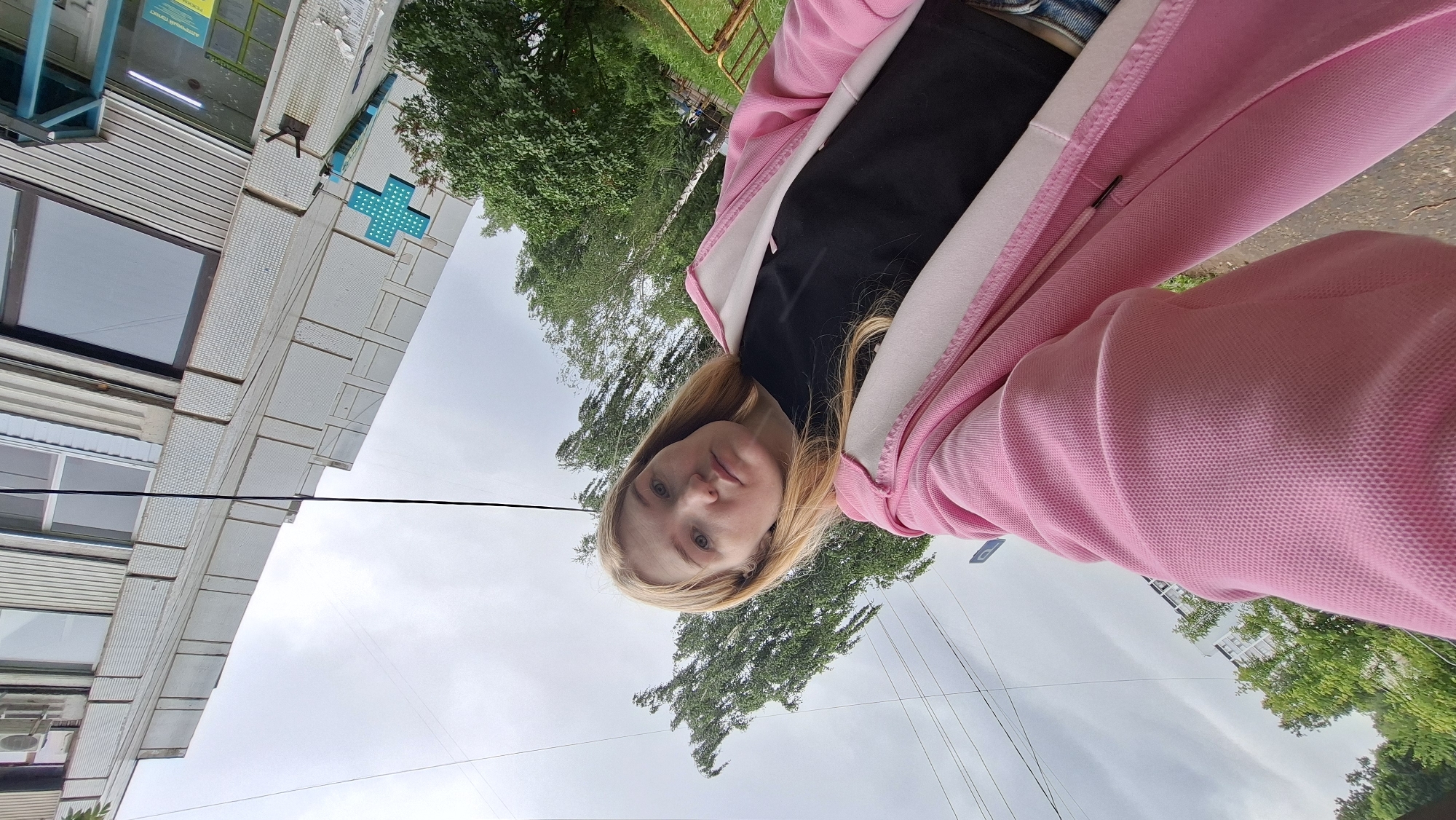 An outdoor, low-angle, medium shot shows a young Caucasian woman with long blonde hair facing forward while standing outside a building in Chaikovsky, Russia. She is wearing a black t-shirt and a pink zip-up hooded jacket. The building behind her is light gray with rows of small, square tiles. A turquoise, dot-patterned cross, a common pharmacy symbol, is visible on the side of the building. Trees with green leaves surround the building. The sky is overcast and gray. Power lines are visible crisscrossing the sky in the background. The camera is positioned at a low angle, looking up at the woman and the building.