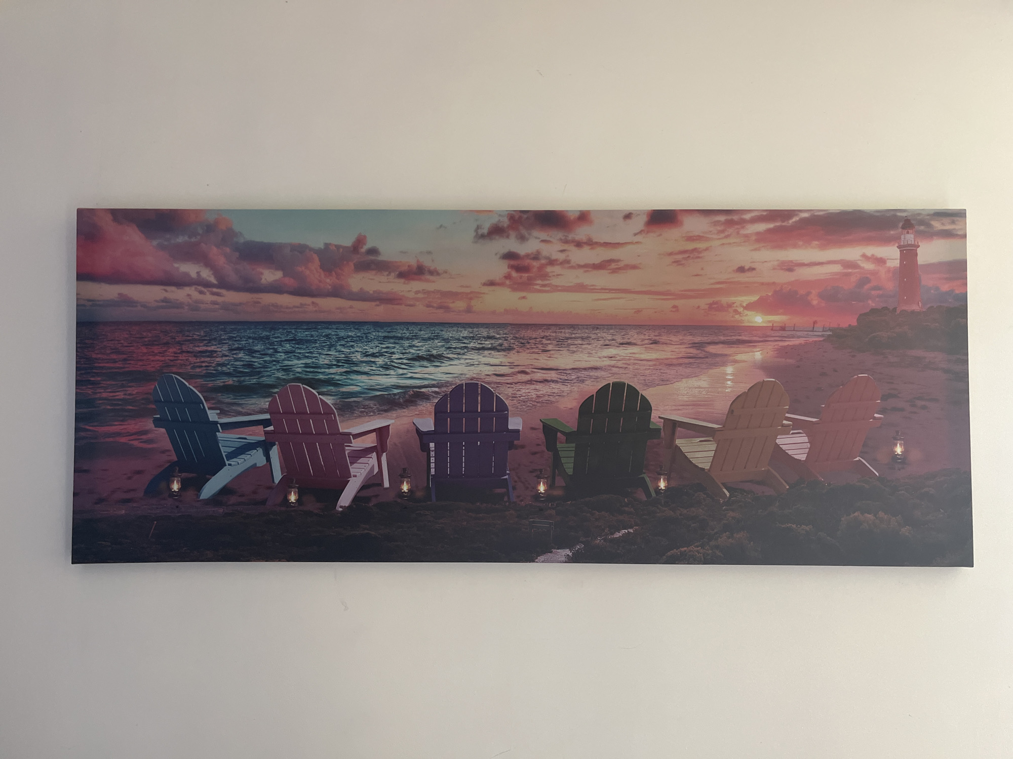 The image depicts a rectangular canvas print hanging on a white wall. The artwork shows a beach scene at sunset with six Adirondack chairs in a row facing the ocean. From left to right, the chairs are colored blue, pink, purple, green, tan, and red. Small lanterns are placed in front of each chair. The sky is filled with pink and purple clouds. A lighthouse stands on a land formation to the right of the frame, near the sun on the horizon. The location is not specified in the image and is therefore not possible to determine.