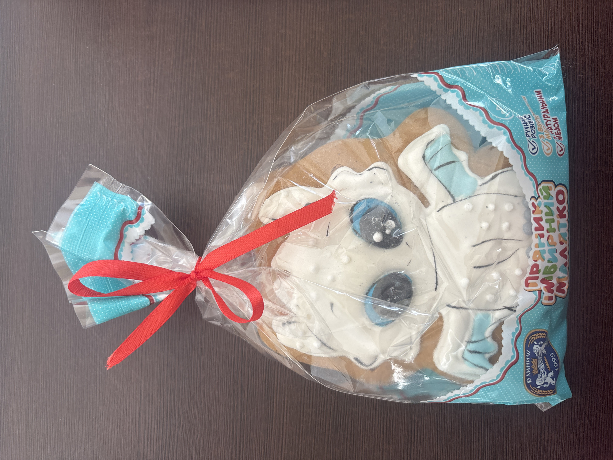 A close-up, top-down shot shows a packaged gingerbread cookie shaped like a cartoon dragon, likely from the animated movie franchise "How to Train Your Dragon". The cookie is decorated with white icing, featuring blue eyes and light blue wings. It is enclosed in clear plastic packaging, secured at the top with a red ribbon tied in a bow. The packaging features a blue polka-dot design and text in Cyrillic script. The bottom left corner of the packaging displays a blue and gold crest with the word "КУЛІНІЧІ" and the year "1995" printed below it, indicating the brand "Kulīnichi" and its founding year. This brand is associated with Kremenchuk, Ukraine. The cookie and packaging are placed on a dark brown wooden surface, likely a table.