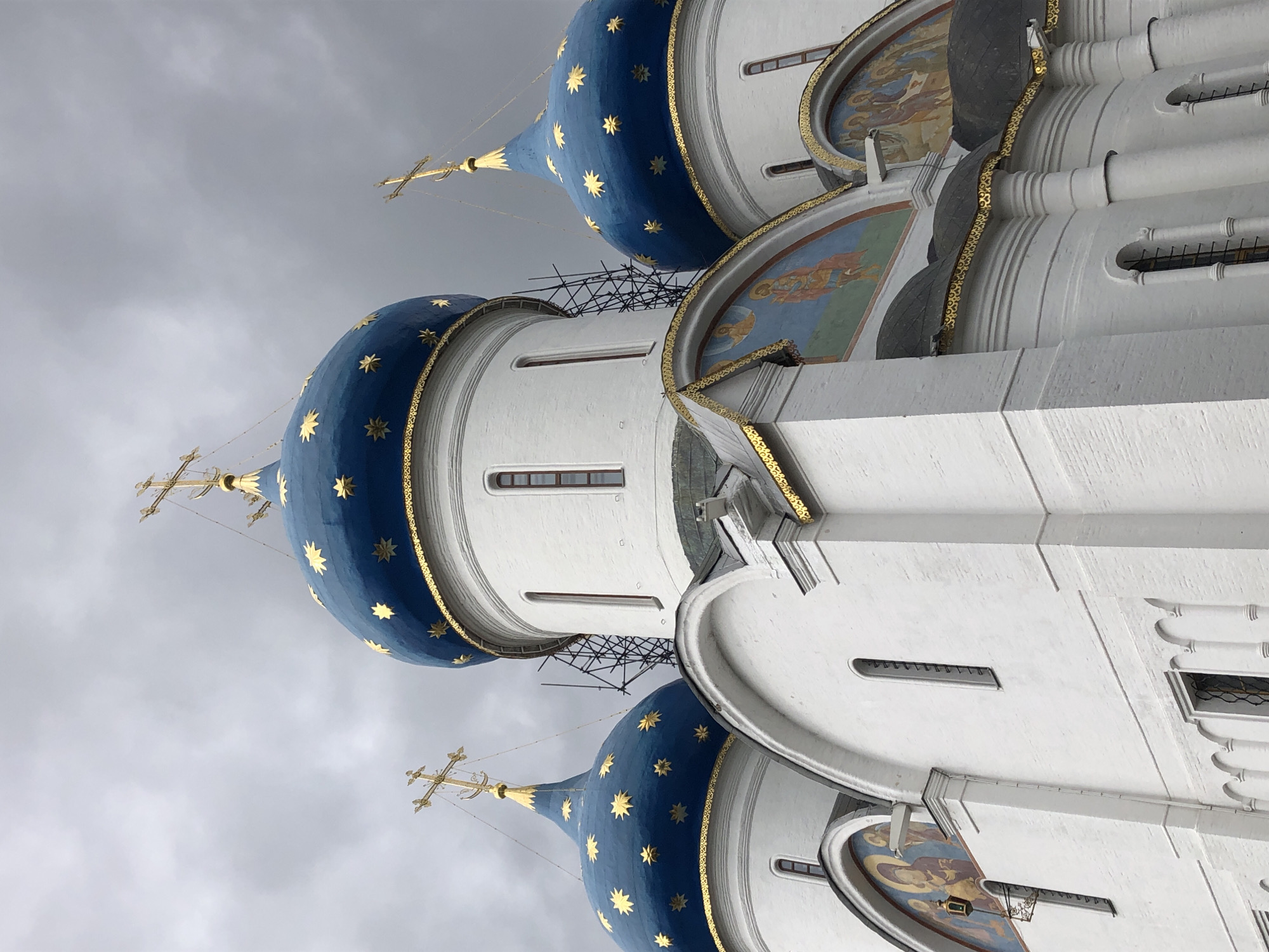 A low-angle, full shot captures the stunning architecture of a white brick church against a dramatic, overcast sky. Three prominent blue domes, each adorned with golden stars and topped with a golden cross, dominate the upper portion of the image. The domes are circular and taper to a point, with a decorative golden band encircling their base. Between two of the domes, scaffolding is visible, suggesting ongoing construction or maintenance. The lower part of the church features white brick walls with arched windows and decorative painted frescoes above some of the arched openings. The frescoes depict religious figures in muted tones. The overall impression is one of grandeur and solemnity, enhanced by the moody lighting of the cloudy sky.