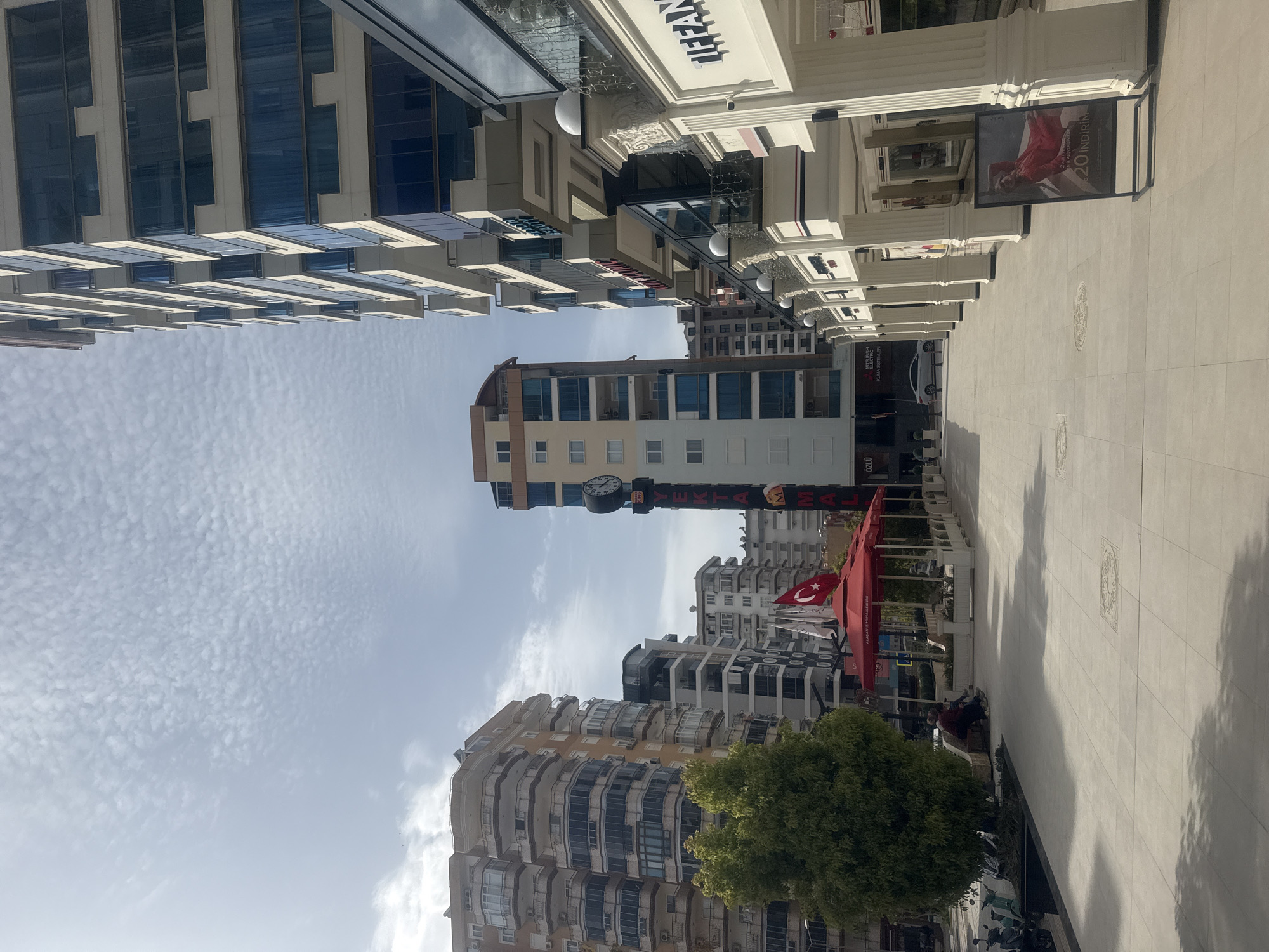 The image captures a street scene in an urban environment. On the left, a tall building with multiple stories and blue-tinted windows reaches towards the sky. The sky above is a pale blue with some clouds. A multi-story building with a clock tower is centered in the shot. To the right, a paved walkway bordered by shops with ornate facades and architectural columns stretches into the distance. A promotional banner and advertisement are displayed on the right, with a person posing for the photograph. A Turkish flag is planted near the red canopies of outdoor seating areas. A green tree is visible in the lower part of the frame. Bicycles and people populate the lower right corner of the image.