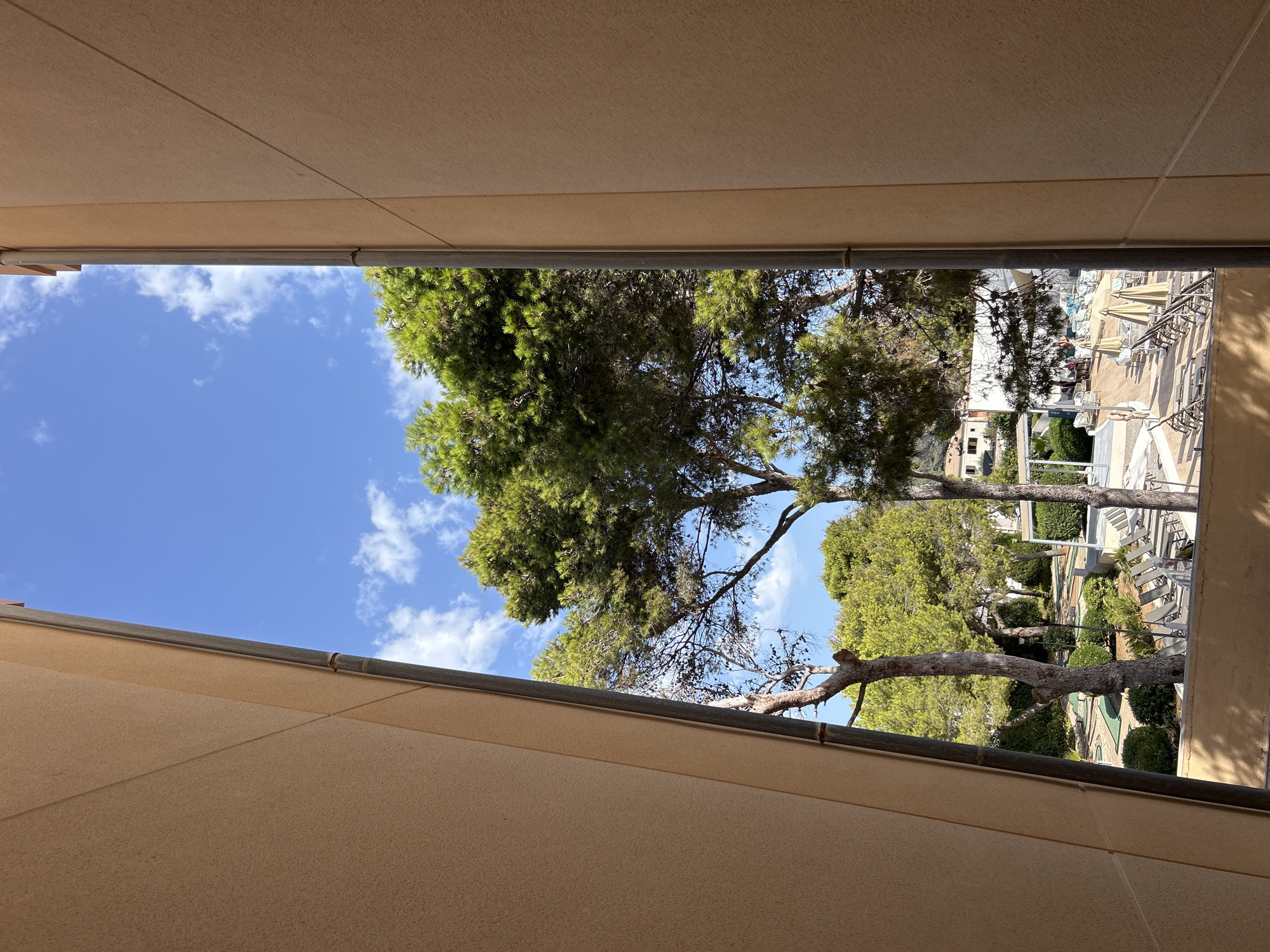 The image shows a view looking out from between two tan building structures. The sky is blue with scattered white clouds. In the distance, there are tall green trees with exposed trunks and branches. Below the trees, there is a resort area with lounge chairs, umbrellas, and what appears to be a miniature golf course. A few people are visible in the resort area. The buildings frame the view, creating a tunnel-like perspective. The environment suggests a sunny day at a vacation resort.