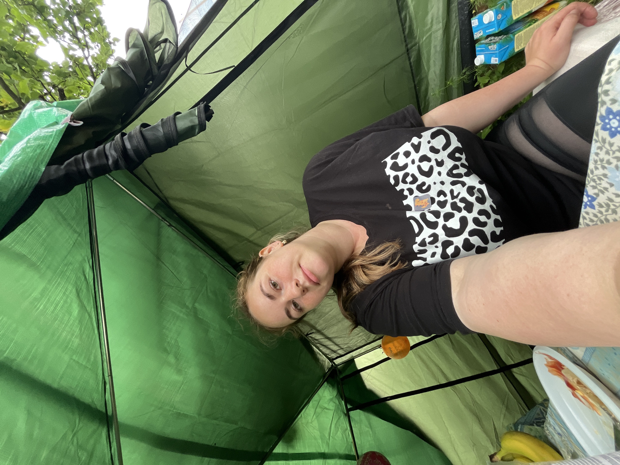 A young woman is taking a selfie inside a green tent. She is wearing a black t-shirt with a white leopard print design on it. Her hair is blonde and she is smiling at the camera. The tent appears to be set up outdoors, with trees and foliage visible in the background. There are also some cartons of juice and a plate with food on a table next to her. The overall mood of the image is relaxed and informal. The lighting suggests it is daytime. No text is clearly legible.