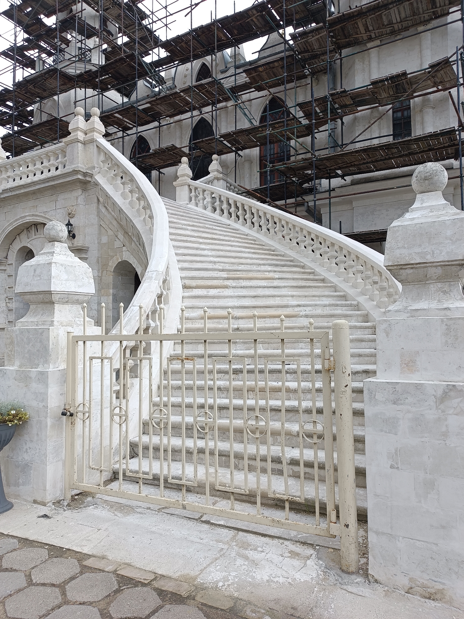 The image shows the grand white stone staircase leading to the Church of the Vladimir Icon of the Mother of God in Bykovo, Russia. The church is currently undergoing extensive renovation, as evidenced by the scaffolding covering its exterior. The staircase itself is a prominent architectural feature, with a sweeping curve and ornate balustrade. A decorative metal gate is positioned at the bottom of the stairs. The ground in front of the gate is paved with hexagonal stones. The overall scene suggests ongoing restoration work on a historic religious building.