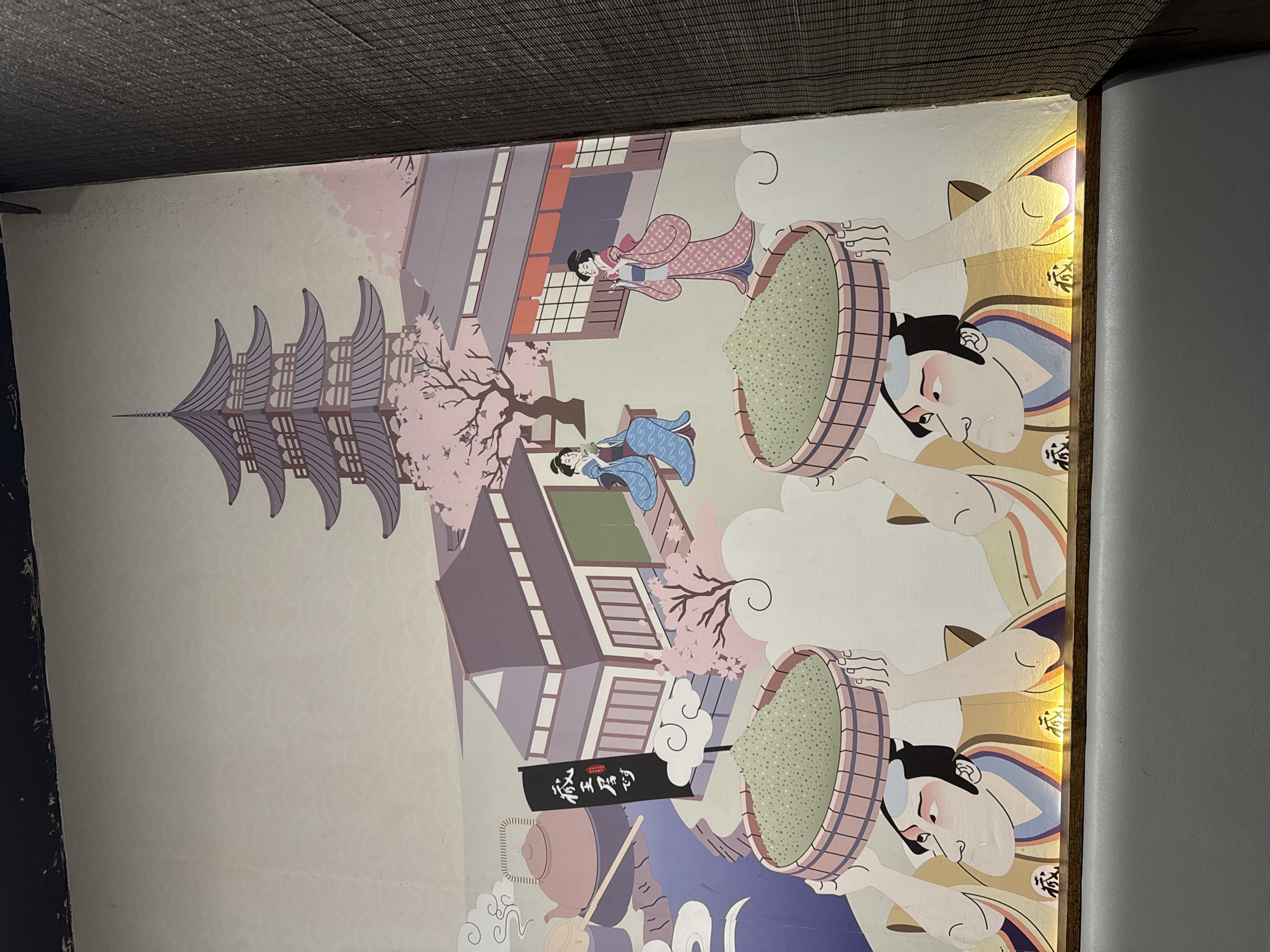 The image shows a mural depicting a traditional Japanese scene, likely a restaurant or cultural establishment. In the foreground, two stylized male figures with exaggerated features are carrying large baskets of green powder on their heads. The baskets appear to be made of woven wood. Behind them, two women in traditional Japanese kimonos are depicted. One woman, seated on a bench, is holding a cup. The other woman is standing. In the background, there are traditional Japanese buildings with tiled roofs and shoji screens, a multi-tiered pagoda, and cherry blossom trees in bloom. A black banner with Japanese characters is also visible. The mural has a light, pastel color palette with accents of pink, purple, and green. The overall style is illustrative and somewhat cartoonish, reminiscent of Japanese woodblock prints. The bottom of the mural is illuminated by yellow lights, suggesting it is mounted on a wall and lit from below. The mural is likely located in New York City, United States of America, given the context of a business or establishment that would commission such artwork. However, without further information, the specific location within New York City cannot be determined.