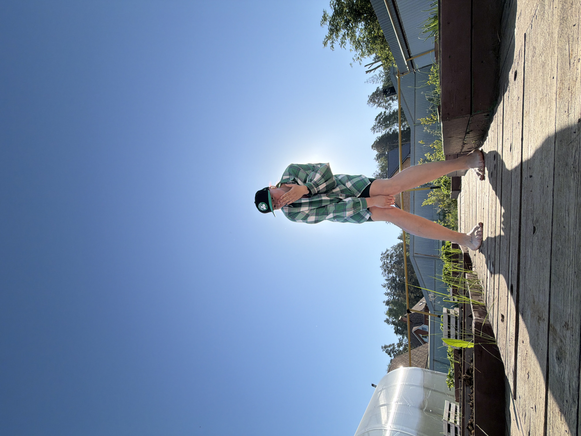 A person is standing barefoot on a wooden platform, backlit by the sun in Chaikovsky, Russia. The individual is wearing a green and white plaid shirt, black shorts, and a black baseball cap with a green logo. Their hands are near their face. The background includes a clear blue sky, trees, buildings, and a greenhouse.