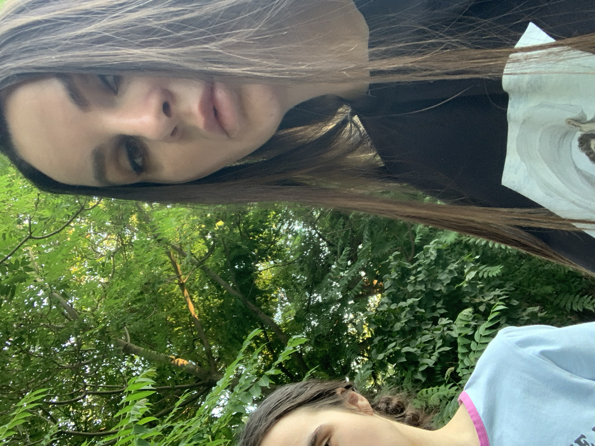 The image is an upside-down, close-up selfie taken outdoors. Two individuals are partially visible. The foreground features the face of a person with long, dark hair, looking directly at the camera. Their features are soft, and their lips are slightly parted. The background is a lush, green canopy of trees, suggesting a natural, possibly wooded setting. Sunlight filters through the leaves, creating dappled light.
The bottom portion of the image shows another person from the shoulders up, also wearing a light blue t-shirt with a pink trim. Their head is turned away from the camera, and only a portion of their face and hair is visible.
The overall impression is casual and relaxed, possibly a moment captured during a day spent outdoors, perhaps in a park or forest. The time of day appears to be daytime, judging by the natural light. There is no visible text in the image.