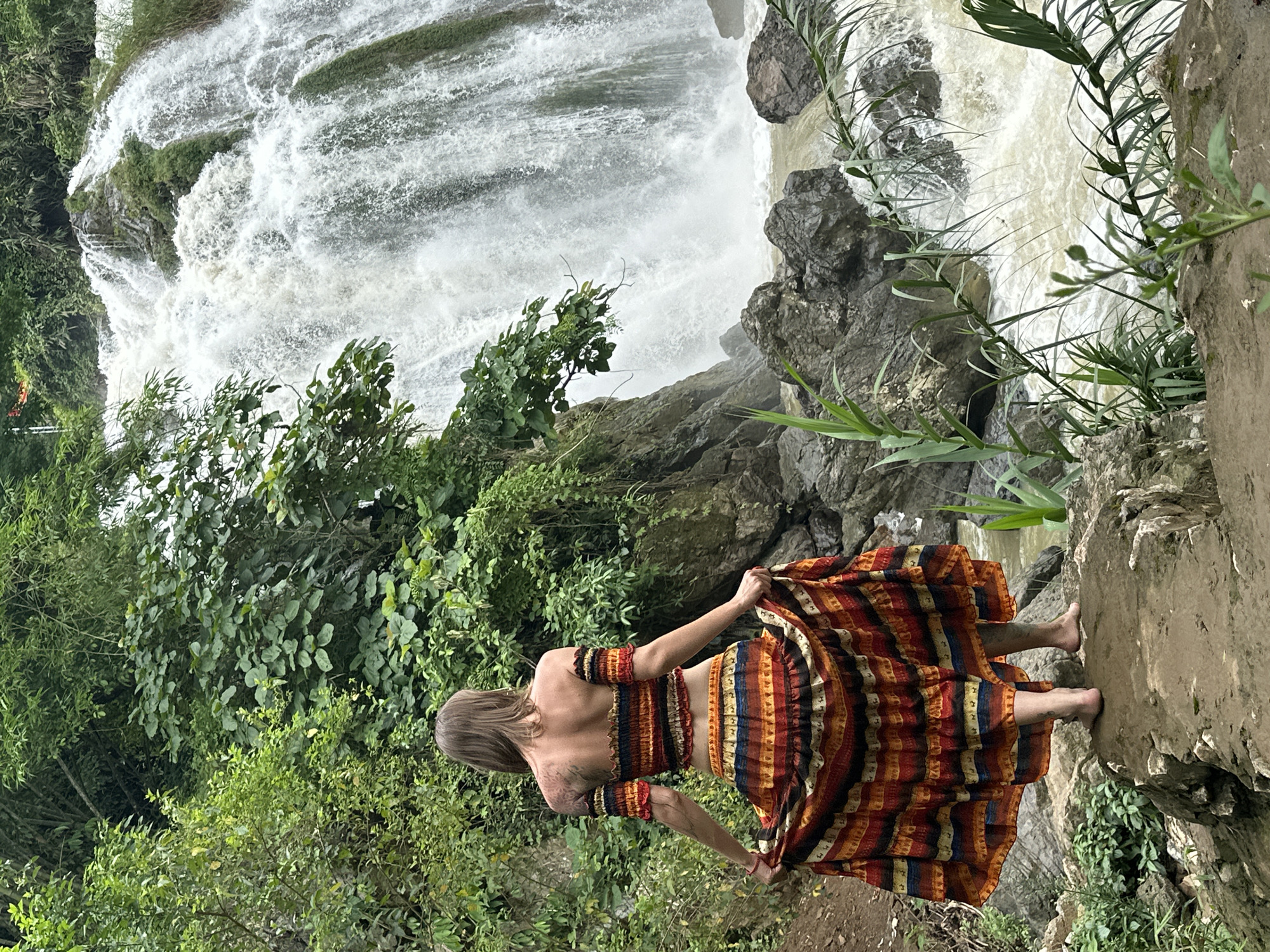 A woman stands barefoot on a rock, facing away from the camera, her hands holding the edges of a flowing, multi-colored striped skirt. She wears a matching off-the-shoulder crop top. Behind her, a powerful waterfall cascades down a rocky cliff face amidst dense green foliage. The scene is located in the vicinity of the Bản Phúc waterfall in Bản Phúc, Vietnam. The water is turbulent at the base of the falls and flows into a river. Rocks and vegetation surround the area.