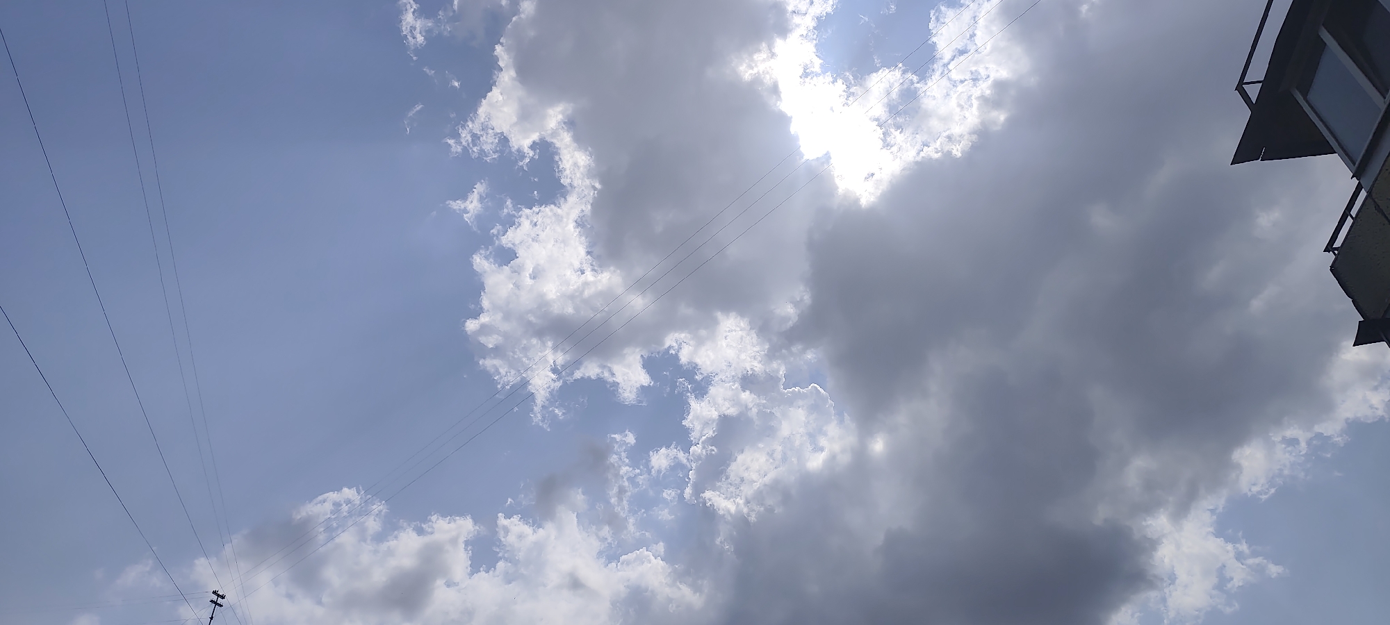 The image captures a view of the sky on a cloudy day with the sun partially obscured by clouds. Several power lines are visible, running diagonally across the frame. In the upper right corner, a portion of a building with a balcony and window is visible. The scene is likely an outdoor urban setting, potentially in Kyiv, Ukraine, given the context of the request, though specific landmarks are not identifiable.