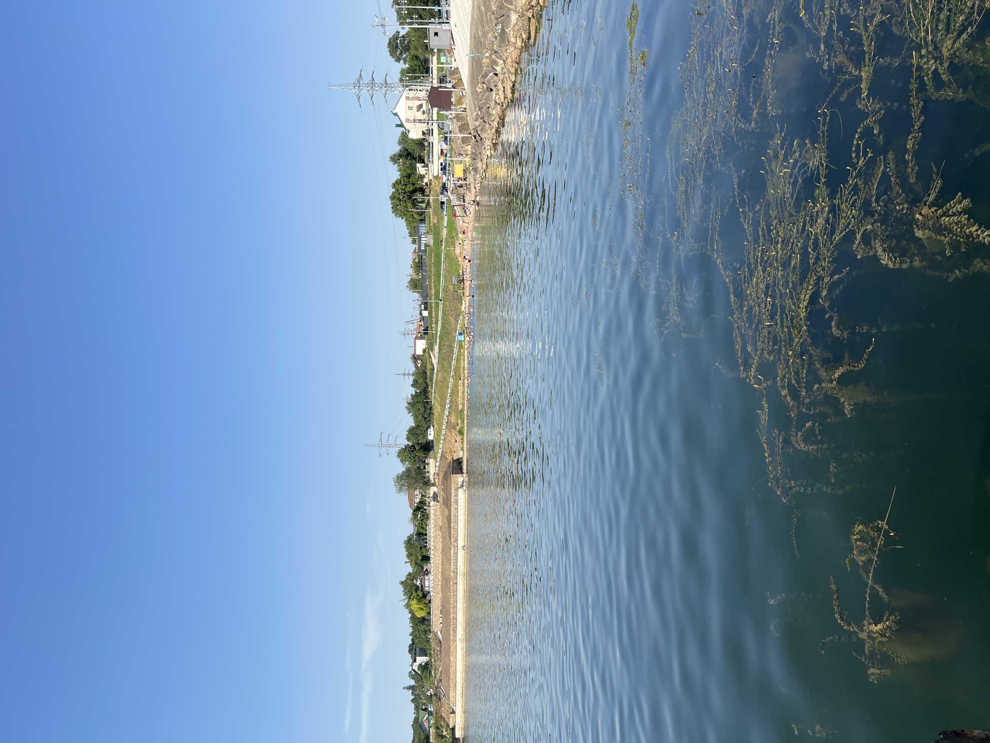 This is a medium shot of a lake on a clear day. The water is dark blue with some ripples and aquatic plants visible near the surface. In the background, there is a beach with many people swimming and sunbathing. Beyond the beach, there is a hill with trees and houses. Power lines and pylons are visible in the distance, indicating a developed area. The sky is clear and blue with no clouds. The location is identified as the city of Korzhevo, Moldova.
