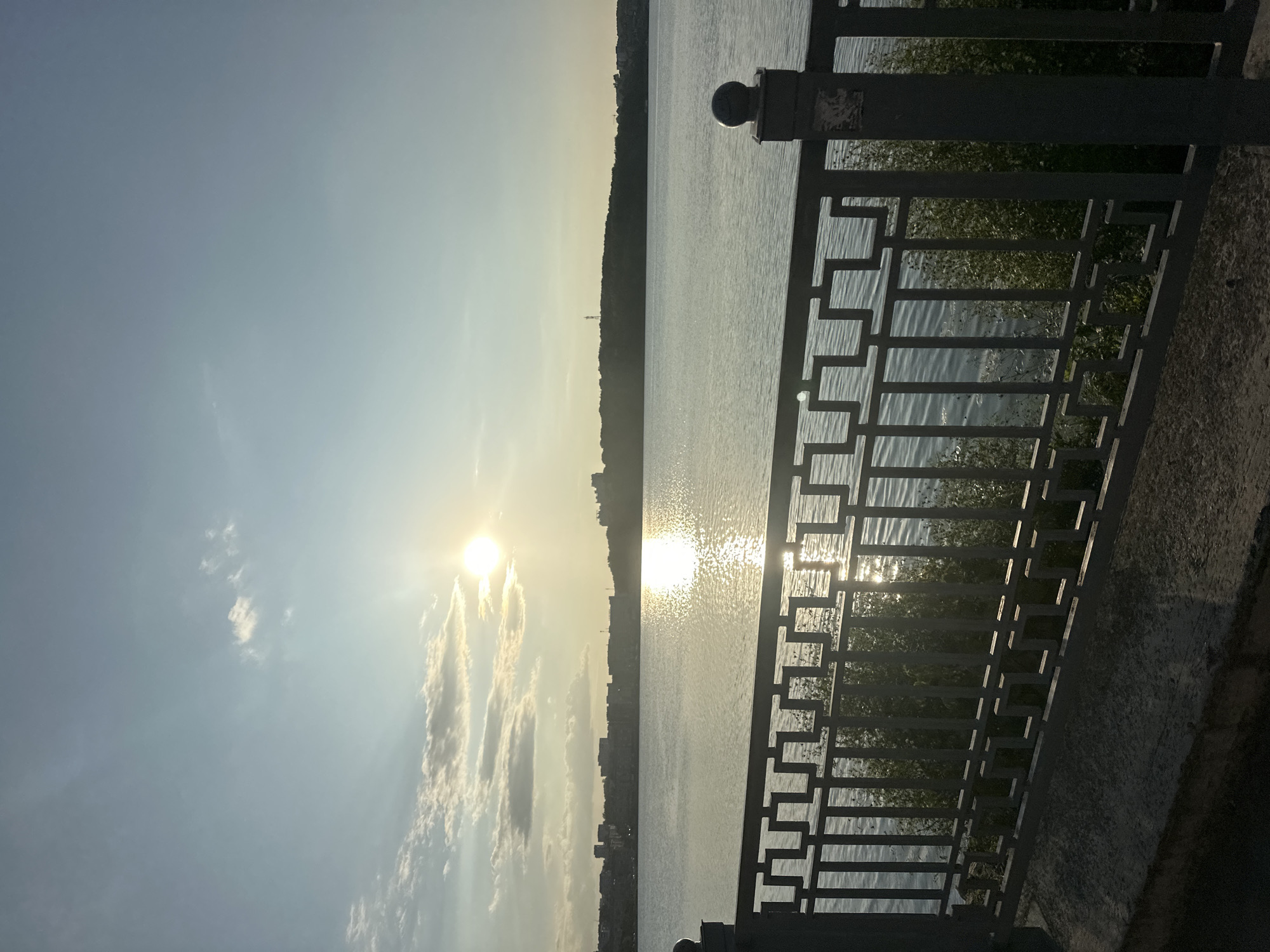 The image depicts a serene waterscape with a city skyline in the distance, under a bright, late afternoon sun. The foreground features a dark metal railing with a geometric pattern, separating a concrete embankment from the body of water. This embankment appears to be a pedestrian walkway or promenade. Beyond the railing, the water reflects the sunlight, creating a shimmering effect. Across the water, a densely built urban area is visible on a distant shoreline, characterized by a mix of buildings and some greenery. The sky is a pale blue with scattered white clouds, and the sun is a bright orb positioned in the upper left quadrant of the sky, casting a strong glare on the water. The overall impression is one of a peaceful urban waterfront during a sunny day. This scene is consistent with views along the Ternopil Reservoir in Ternopil, Ukraine, a popular waterfront area offering panoramic views of the city and surrounding landscape.