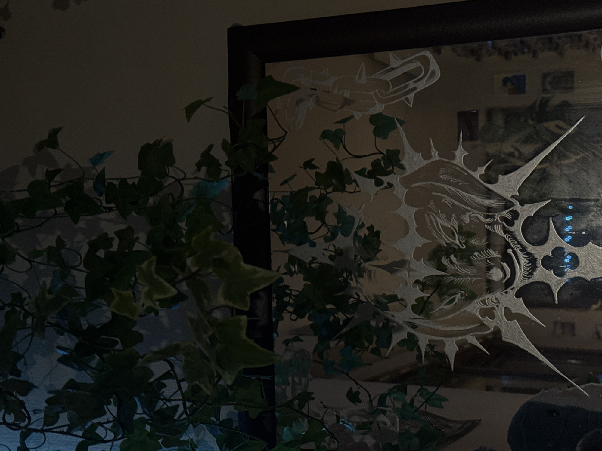 The image shows a close-up of an ornate mirror, framed in dark wood. The mirror's surface is etched with intricate designs, including what appears to be a stylized demonic face with sharp, pointed elements radiating outwards, and another figure on the right that resembles a chained or bound entity with spikes. In the foreground, a dark green ivy plant with some lighter-colored leaves hangs down over the top of the mirror, partially obscuring the reflection. The reflection in the mirror shows a dimly lit room with other objects visible, including what seems to be framed artwork on a wall. The lighting is dim and atmospheric, with some blueish light contributing to the overall mood. The scene is indoors, likely in a residential or decorative setting. The location in the city of Cork, Ireland, is not discernible from the image.