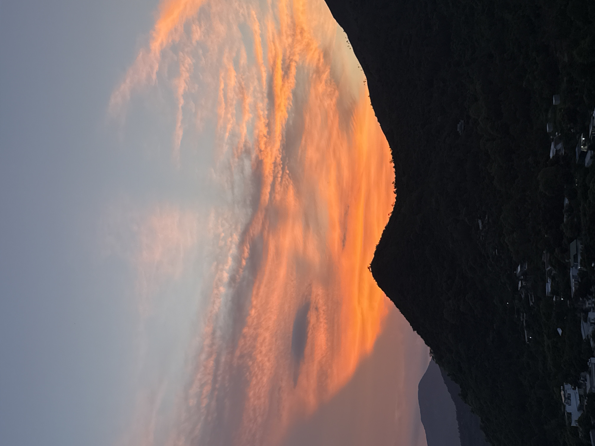 The image captures a dramatic sunset over a mountainous landscape with a settlement nestled on the slopes. The sky is dominated by vibrant orange and pink clouds, illuminated by the setting sun, creating a warm and ethereal glow. The clouds are streaky and wispy, adding texture and depth to the scene.
On the right side of the image, a dark, forested mountain slopes down towards a cluster of buildings. The settlement appears to be illuminated with lights, suggesting it's evening. The architecture of the buildings is not clearly visible due to the dark shadow and distance. In the distance, more mountains can be seen, appearing as silhouettes against the colorful sky.
The overall mood is one of tranquility and natural beauty, with the intense colors of the sunset contrasting sharply with the dark, imposing mountains. The image evokes a sense of peacefulness and wonder at the spectacle of nature. The time of day is clearly sunset, given the vibrant colors in the sky. The location is identified as Bac Nha Trang Ward, Vietnam, which aligns with a tropical coastal mountainous setting. No text is visible in the image.