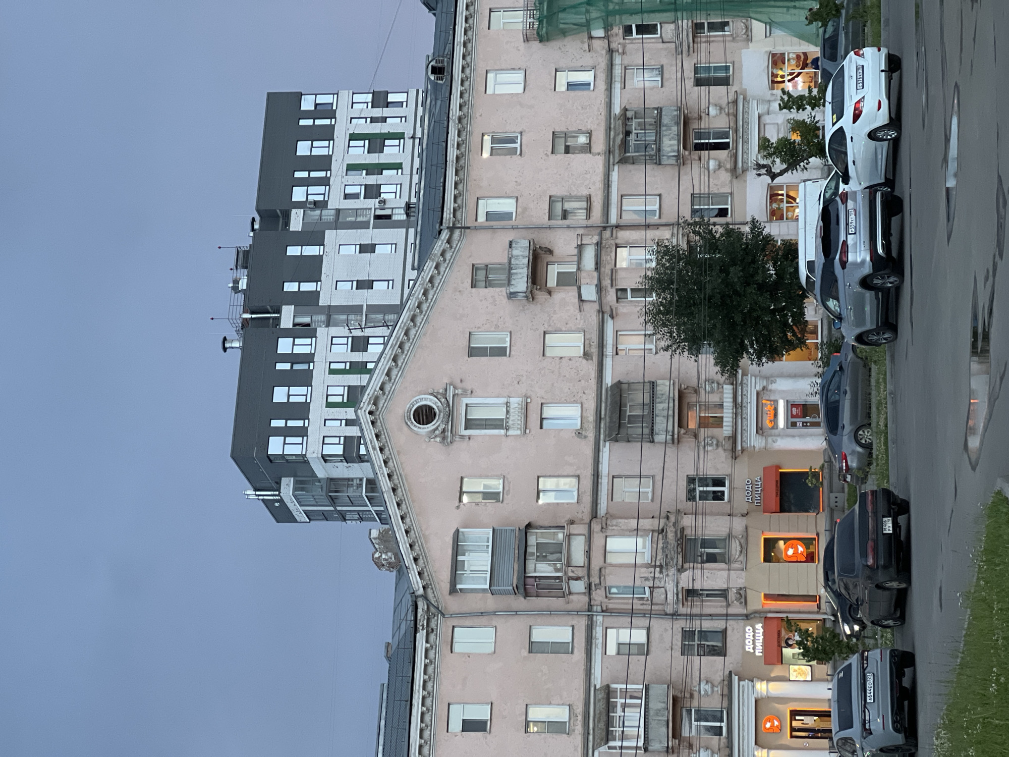 The image depicts a street scene in Izhevsk, Russia, with buildings and parked cars. Two apartment buildings are visible. The building on the left is modern and features a grey and white facade with numerous windows arranged in a grid pattern. The building on the right is older, with a pastel pink, weathered facade and a more complex architectural design, including a pointed roof section with a circular window. Several cars are parked along the street in front of the older building. These include a white sedan, a grey SUV, and two black cars. Some of these cars have visible license plates.
Several commercial establishments with illuminated signage are located on the ground floor of the older building. Signs such as "Dodo Pizza" (in Russian: "Додо Пицца") are clearly legible, along with orange glowing circular logos associated with the pizza chain. Other storefronts are also visible, with varying degrees of illumination.
Overhead, power lines crisscross the scene. Trees with green foliage are present, suggesting a period outside of winter. The sky is overcast and grey, indicating it is likely late afternoon or early evening, and the lighting suggests it is twilight. There are no people visibly present in the image. The overall impression is a typical urban streetscape in a Russian city.