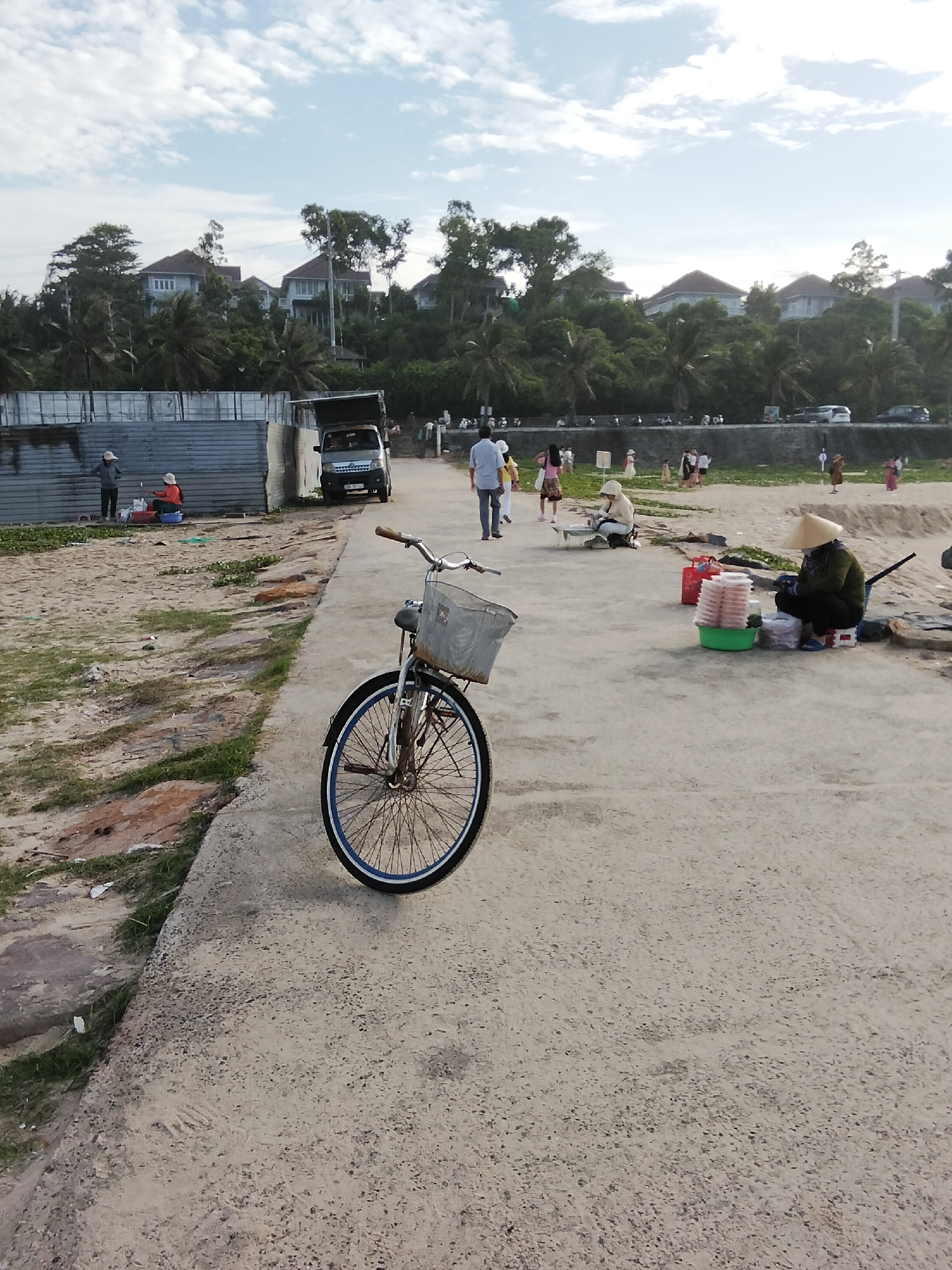 A bicycle with a wire basket is parked on a concrete path. To the left of the path is a sandy area with some sparse vegetation and rocks. In the background, a low, corrugated metal wall separates the sandy area from a street with several parked cars and a small truck. Beyond the street, lush green trees and palm trees are visible, with several modern houses situated on a hill. Further in the background, the sky is a pale blue with scattered white clouds. Several people are seen walking along the street or near the sandy area. In the mid-ground on the right side of the path, a person wearing a conical hat is sitting near a stack of containers filled with food and a red bag. Another person is sitting behind a table on the left side of the path. The scene appears to be a public area near the coast in Phú Thủy, Vietnam.