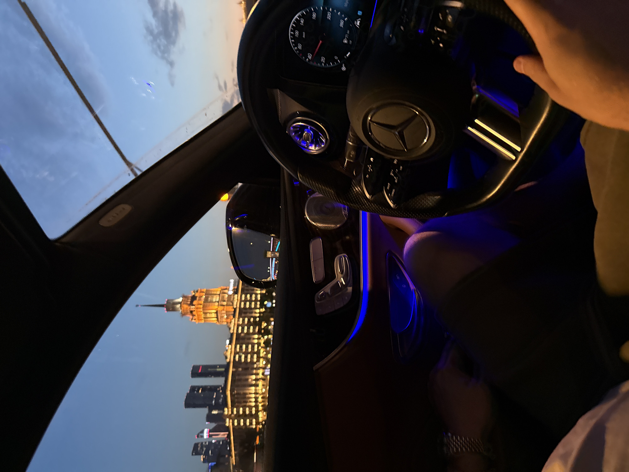 The image is taken from the interior of a car, looking out at a cityscape at dusk. The sky is a deep blue with some clouds. In the distance, several tall buildings are illuminated, including a prominent, ornate building with a spire that appears to be the Hotel Ukraina in Moscow, Russia. The car's interior is dimly lit, with blue ambient lighting visible on the door panel and around the air vents. A person's hands are visible, one on the steering wheel, which features a Mercedes-Benz emblem, and another resting on their lap. The dashboard displays are illuminated. The overall impression is a view of Moscow from a luxurious vehicle at twilight.