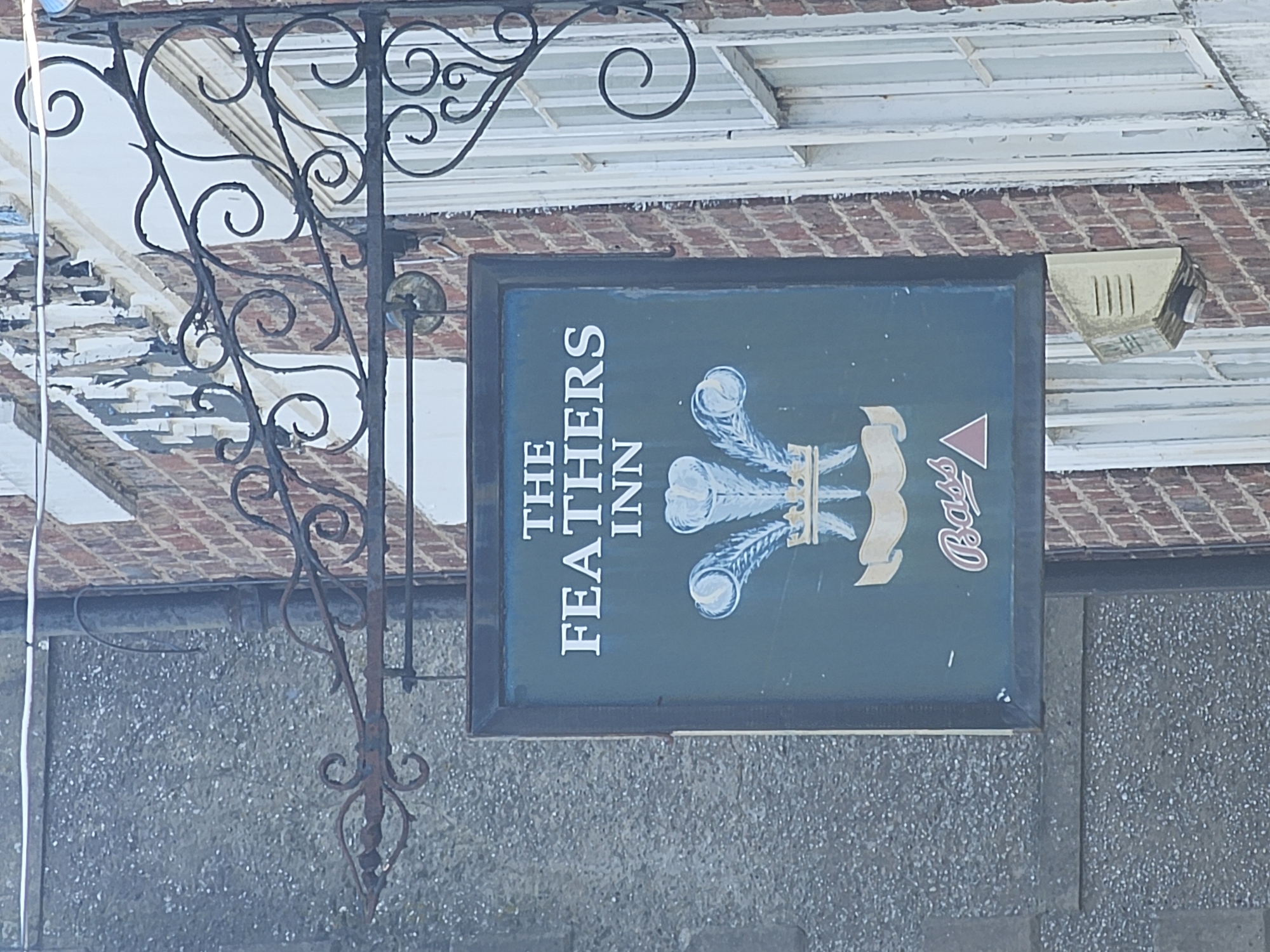 The image shows a pub sign, currently oriented 90 degrees clockwise, suspended from an ornate wrought iron bracket. The sign itself is rectangular with a dark green background, featuring white and gold text and imagery. On the left side, vertically aligned, the words "THE FEATHERS INN" are visible in white uppercase letters. To the right of the text, an illustration of three stylized feathers, possibly ostrich plumes, is depicted in shades of white and light blue with golden accents. Below these feathers and to the right, the distinct red triangular logo of "Bass" brewery is present, also rotated.
The elaborate wrought iron bracket, dark and showing signs of rust, supports the sign from an attachment point on a building. It features numerous decorative scrolls and curls, extending both above and below the sign.
The background reveals sections of a building under clear daylight. Above the sign, an aged brick wall is visible, topped by white wooden window frames or fascia boards that show peeling paint and signs of disrepair. To the right, mounted on the brickwork, is a light-colored, rectangular object with vertical slits, possibly a speaker or vent. Below the sign and the lower part of the bracket, a textured grey wall, likely pebbledash, forms the lower facade of the building.
The scene is well-lit by natural light, indicating it is daytime. The presence of "THE FEATHERS INN" sign and the traditional architecture suggest a public house setting in Holywell, United Kingdom.