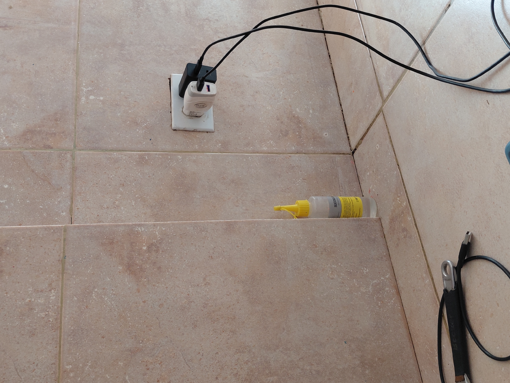 The image displays a ground-level view of an indoor tiled corner, likely in a domestic setting in Santa Ana, Venezuela. The floor and a section of the lower wall are covered in light beige or brownish ceramic tiles with a subtle speckled pattern, separated by lighter grout lines.
In the upper left portion of the frame, a white electrical outlet is visible on the wall. Two power adapters are plugged into it: a black adapter and a white "Dreamy" branded adapter, which is plugged into the black one. Two black charging cables extend from these adapters, trailing across the tiled floor towards the right side of the image.
Further to the right, resting on a tiled surface that appears to be a step or raised section, is a translucent plastic bottle with a yellow pointed nozzle. The label on the bottle is partially visible, showing the words "SILICON" and "SILICONA," indicating it contains silicone sealant or glue. Below and to the right of the bottle, partially visible at the bottom edge of the frame, is a dark-colored tool, possibly a pair of pliers or a crimping tool, with a metallic ring at one end. Another black cable is coiled near this tool.
The scene suggests an area used for charging electronic devices and possibly for household repairs or DIY tasks, given the presence of the silicone and tool. The lighting is consistent, indicating an indoor environment.
