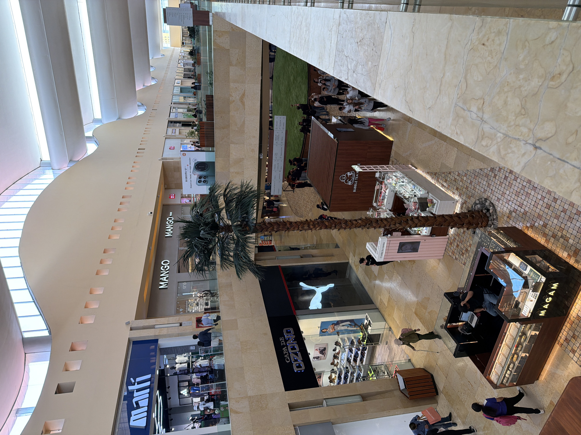 The image, taken from an elevated perspective looking down, captures the interior of a modern, multi-level shopping mall, likely in Puebla, Mexico. The scene is bustling with shoppers and displays, illuminated by bright artificial lighting and possibly natural light from unseen skylights along the high, arched ceiling.
On the ground floor, a large palm tree stands prominently, surrounded by various retail establishments. Visible store names include "MANGO" and "Martí". A store with the text "CAPA DE OZONO" features a large "PUMA" logo prominently displayed in its window. Numerous individuals are seen walking and browsing. Several kiosks are active, one clearly labeled "BUBBLE TEA HO" and another, with a hexagonal shape, displaying "MAGAM". People are seen interacting with these kiosks, and a person is working at the MAGAM stand.
An upper level features a group of people, possibly students or a tour group, gathered near a glass railing overlooking the ground floor. The mall's architecture includes a long, curved wall with evenly spaced square cutouts, and the flooring consists of large, light-colored marble-like tiles and smaller, intricately patterned tiles near the kiosks. A banner advertising "Xiaomi 17" (likely a phone model) with the slogan "en Telcel, mi mejor decisión" is also visible. Other partial text includes "Cuida tu canta" and "MANGO MAN". The mall appears well-maintained and active during daytime hours.