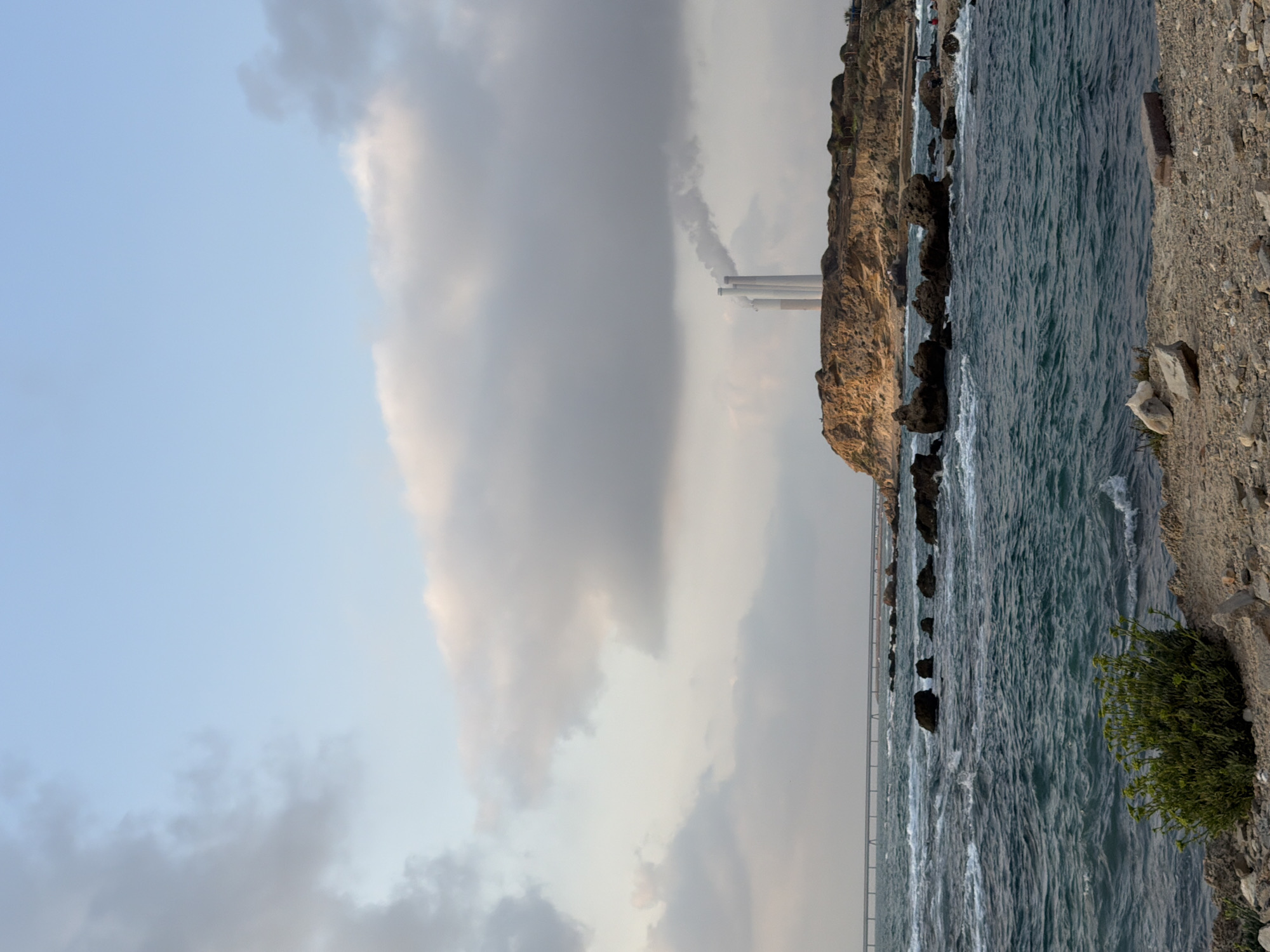 The image shows a coastal scene with a body of water in the foreground and a rocky cliff with industrial smokestacks in the background. The water is choppy with whitecaps, suggesting a windy or wavy condition. On the rocky shore in the foreground, there is a patch of green vegetation and scattered stones. In the distance, a long bridge-like structure extends over the water. The cliff is topped with a power plant with three tall, white smokestacks emitting plumes of smoke, indicating industrial activity. The sky is partly cloudy with a mix of blue and gray clouds. The scene appears to be located near the Orot Rabin power station in Hadera, Israel, based on the presence of the power plant and the coastal landscape. There are a few indistinct figures on the beach in the far distance, appearing to be people.