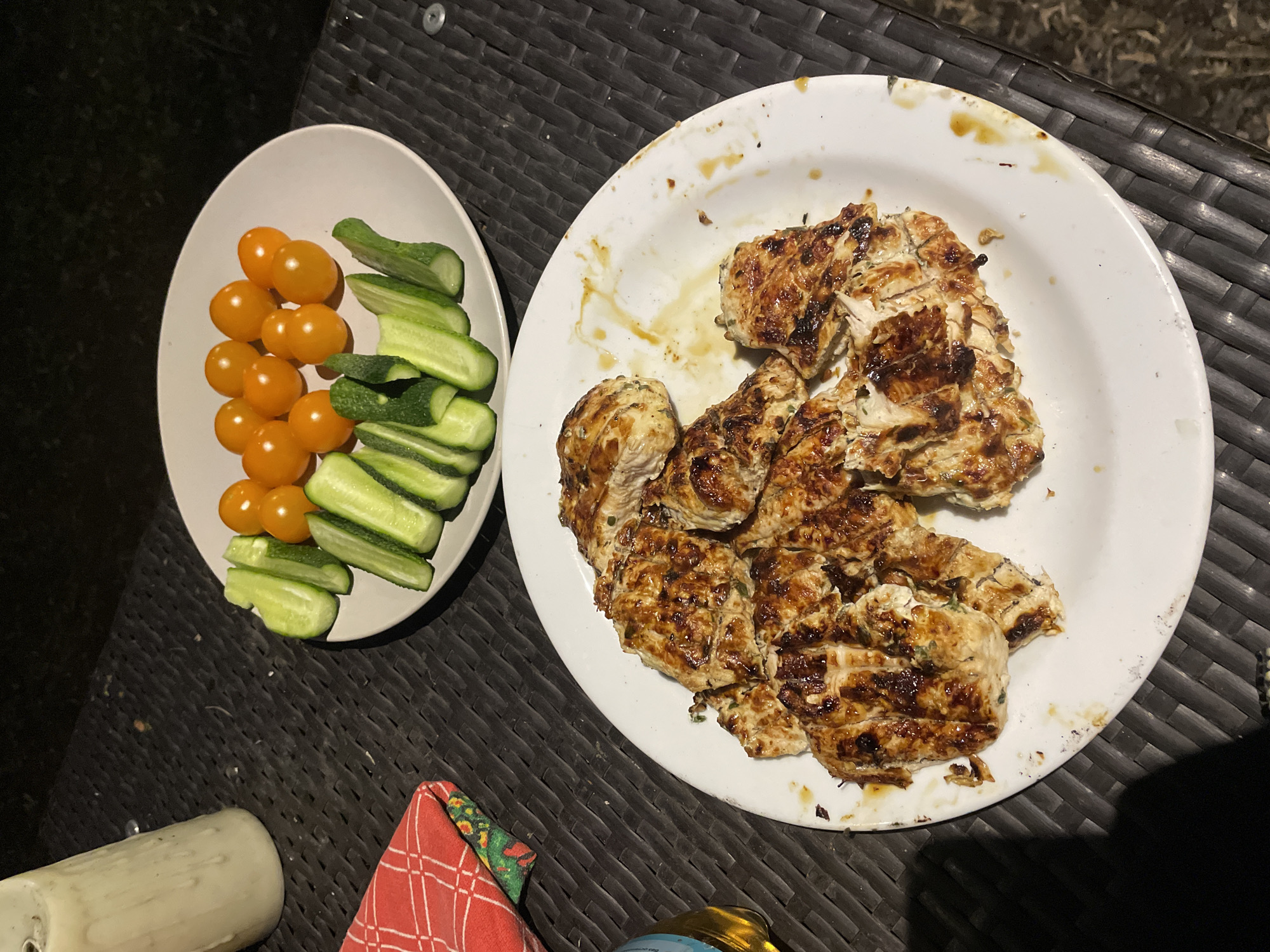 This image presents an overhead view of a meal served outdoors at night. On a dark, woven-patterned outdoor table, a large white circular plate holds numerous pieces of grilled chicken, characterized by their golden-brown char marks and glistening glaze, suggesting a marinade or sauce. Remnants of sauce or cooking residue are visible on the plate.
Beside the grilled chicken, an oval white bowl contains a vibrant side dish of fresh vegetables: a generous arrangement of bright yellow cherry tomatoes and uniformly sliced green cucumbers.
The dark background indicates that the photograph was taken at night or in a dimly lit outdoor setting. The table's texture and visible silver screw heads confirm its outdoor furniture nature. In the bottom left, a white cylindrical object is partially visible, while a red and white checkered fabric item with green and orange patterns, possibly a napkin, is seen below the plates. A small, golden-hued object is also partially visible at the bottom right.
The scene depicts an outdoor dining event, likely dinner, with no people visible. The context suggests the location is Moscow, Russia, though no specific visual cues within the image confirm this. There is no visible text in the image.