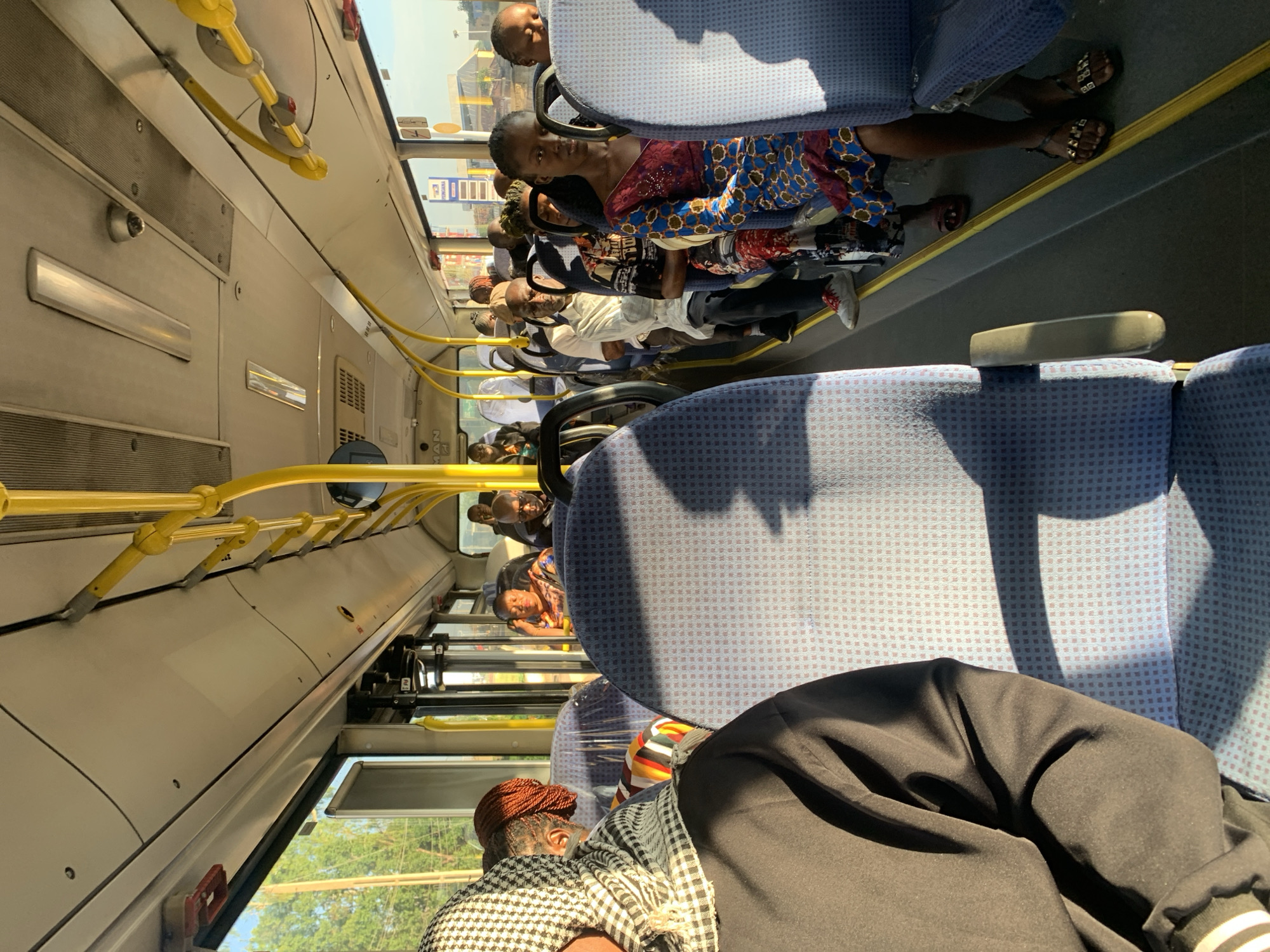 The image shows the interior of a crowded public bus, likely a commercial bus service operating within or to Rafin Par, Nigeria, given the context provided. Sunlight streams in through the windows, illuminating dust motes and casting long shadows across the aisle and seats. The seats are upholstered in a blue patterned fabric, and yellow support poles run along the ceiling and aisle.
On the left side of the bus, in the foreground, a person wearing a black garment and a black and white checkered headscarf is seated, facing towards the front of the bus. Their right shoulder and arm are visible. To their right, in the next row, another person with reddish-brown braided hair is seated, also facing forward.
Further down the aisle, several passengers are visible. A woman in a colorful patterned dress is seated on the right side, looking towards the camera. In front of her, a man in a white shirt and dark trousers is seated. Other passengers, mostly obscured, occupy the seats stretching towards the back of the bus. The overall atmosphere suggests a typical journey during daylight hours, with passengers appearing to be engaged in their own thoughts or conversations, typical of public transportation. The scene is captured from a low angle, emphasizing the perspective of someone seated within the bus.