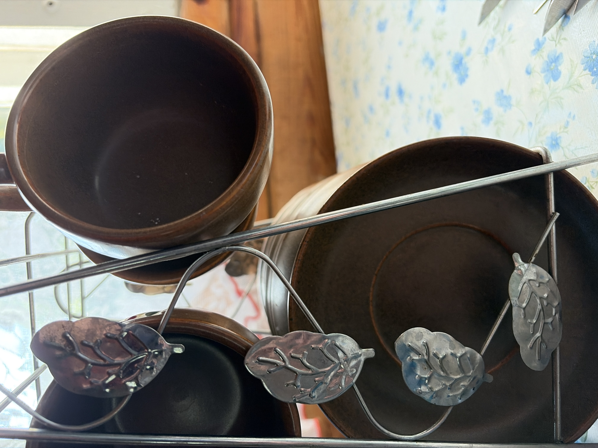 The image shows a close-up of a metal rack holding several dark brown ceramic mugs and saucers. The rack is made of thin metal rods and features decorative silver leaf motifs attached to the supports. Two mugs are visible, one is mostly empty and facing away, while another is partially visible on the left. Three saucers are also present. The rack is placed on a table covered with a white tablecloth featuring a pattern of small blue flowers. In the background, there is a wooden surface, possibly a table or shelf, and a window with a grid pattern, suggesting an indoor setting. The lighting is natural, coming from the window, and casts soft shadows. There are no people visible in the image. The scene does not contain any elements that would allow for the specific location in Malyshevo, Russia to be determined.
