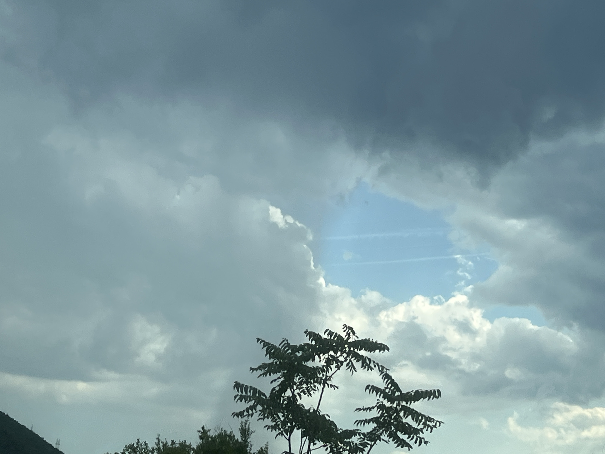 This media file depicts a dramatic sky with heavy, dark clouds obscuring much of the view. A large opening in the clouds reveals a patch of bright blue sky with wispy white clouds and faint contrails, suggesting an airplane passed overhead. In the foreground, the dark silhouette of a leafy tree is visible against the lighter sky. The overall impression is one of an impending storm, with the sun attempting to break through. The lighting suggests it could be late afternoon or early evening. The scene is set outdoors, with hints of hills or mountains on the left side of the frame, suggesting a mountainous coastal region. The dark, brooding clouds evoke a sense of atmospheric tension. There is no visible text in the image.
