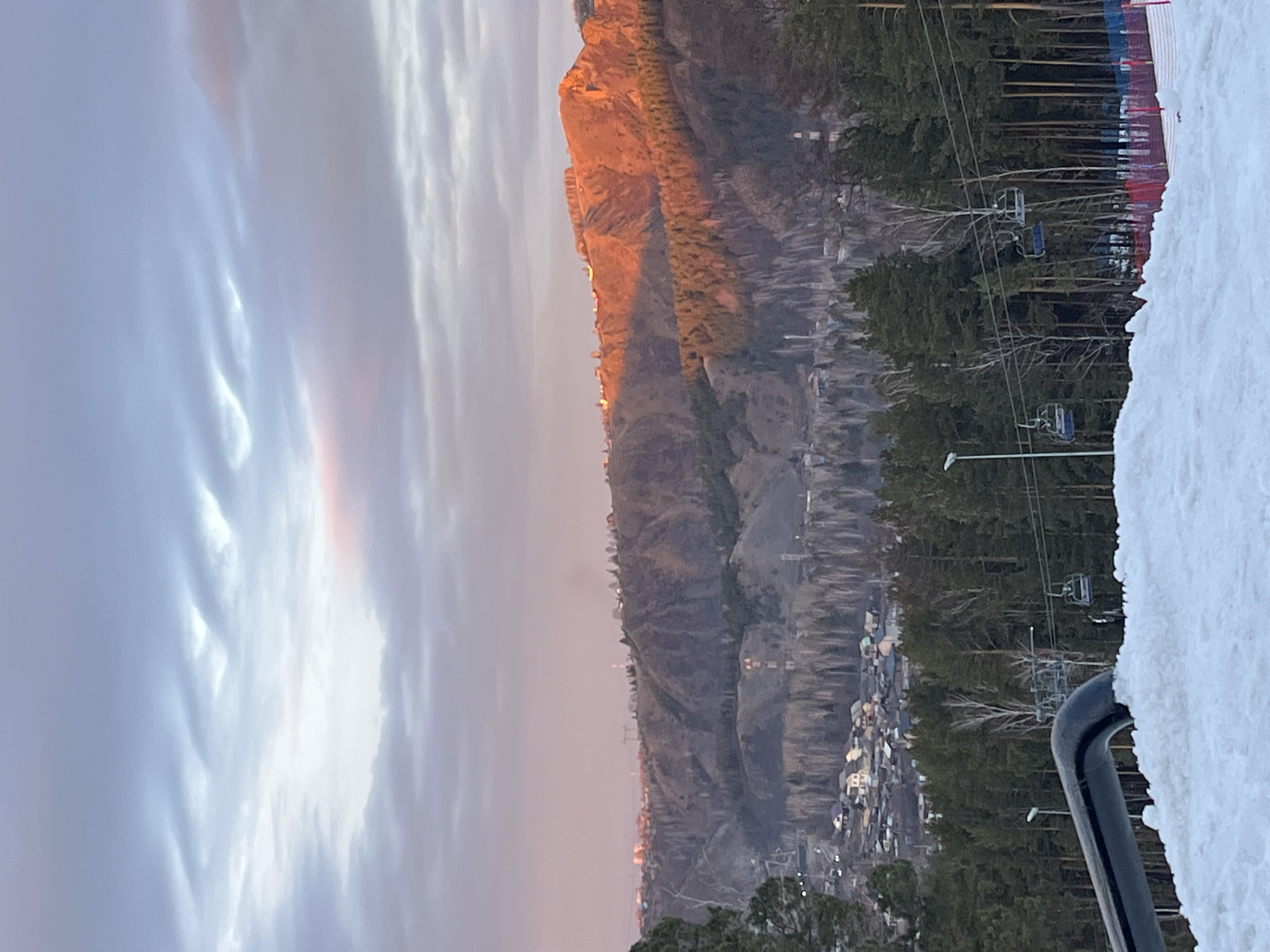 The image depicts a mountainous landscape, likely captured during twilight. To the left of the frame, a clouded sky shows tinges of light orange and purple. Dominating the center is a tall, rocky mountain with a town at its base. A ski lift passes over a forest to the right, adjacent to a snow-covered slope. A railing is in the lower-right corner.